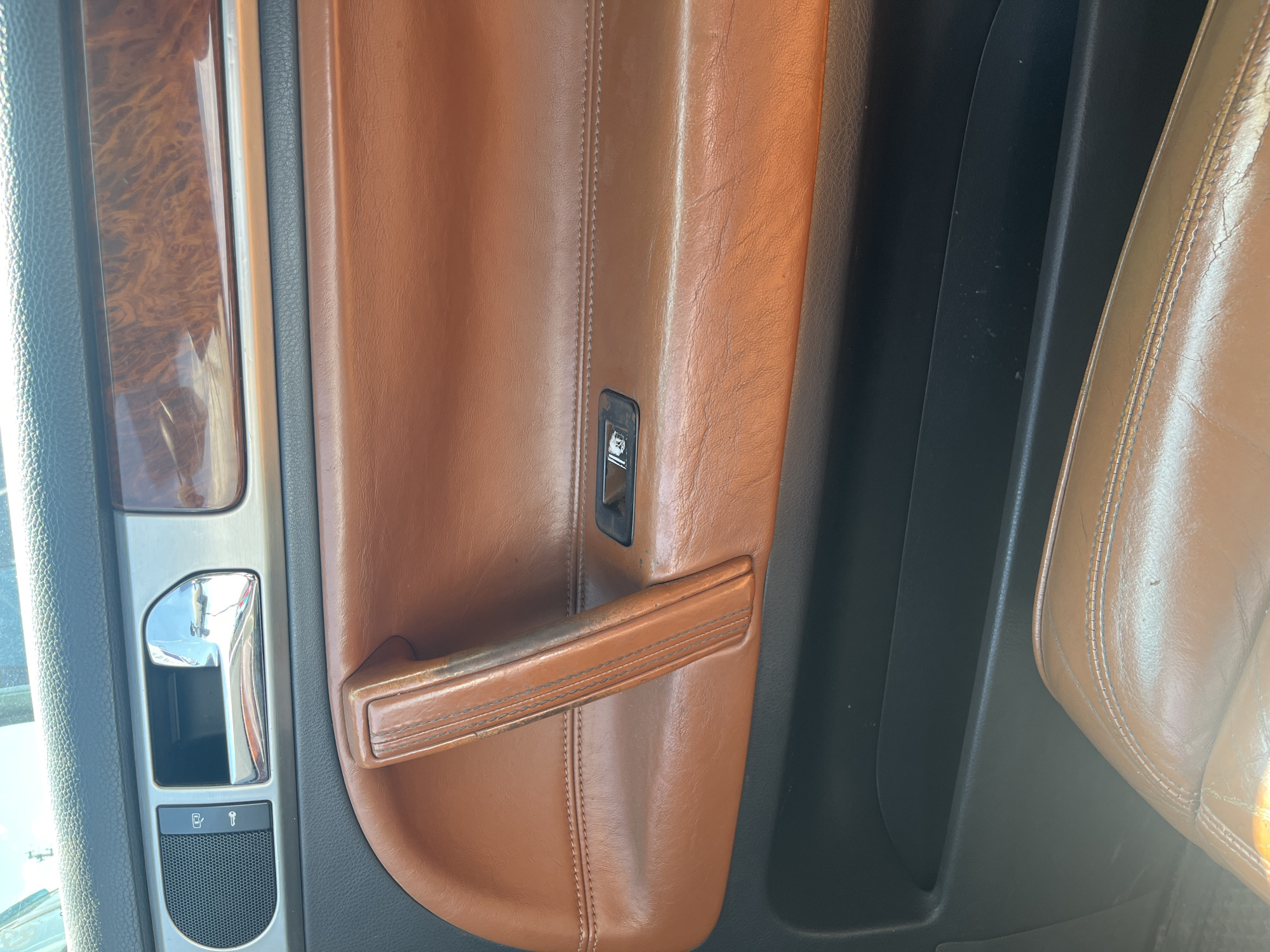 This image shows the interior of a car, likely a luxury model, with a focus on the door panel. The dominant colors are warm browns from the leather upholstery and wood trim, contrasting with the cool metallic finishes of the door handle and controls.
The subjects are the car's interior components. We see a brown leather door panel with prominent stitching, a pull-handle also in brown leather, and a smaller pull tab above it. To the left, there is a section with a wood-grain trim insert, a silver metallic surround, a chrome door handle, and a control panel with buttons and a speaker grille. A single window control button is visible on the brown leather portion of the door. To the right, the edge of a car seat, also upholstered in brown leather, is visible.
There is no activity or event taking place; the image is a still shot of the car's interior.
Notable details include the high-quality materials like leather and wood, suggesting a premium vehicle. The lighting appears to be natural, possibly daylight, casting soft reflections on the metallic surfaces. The location cues are minimal, but the context provided places this car in Aqköl, Kazakhstan. There are no visible emotions conveyed as the image focuses on objects.
There is no visible text in the image, other than the icons on the control panel which are too small to decipher clearly.