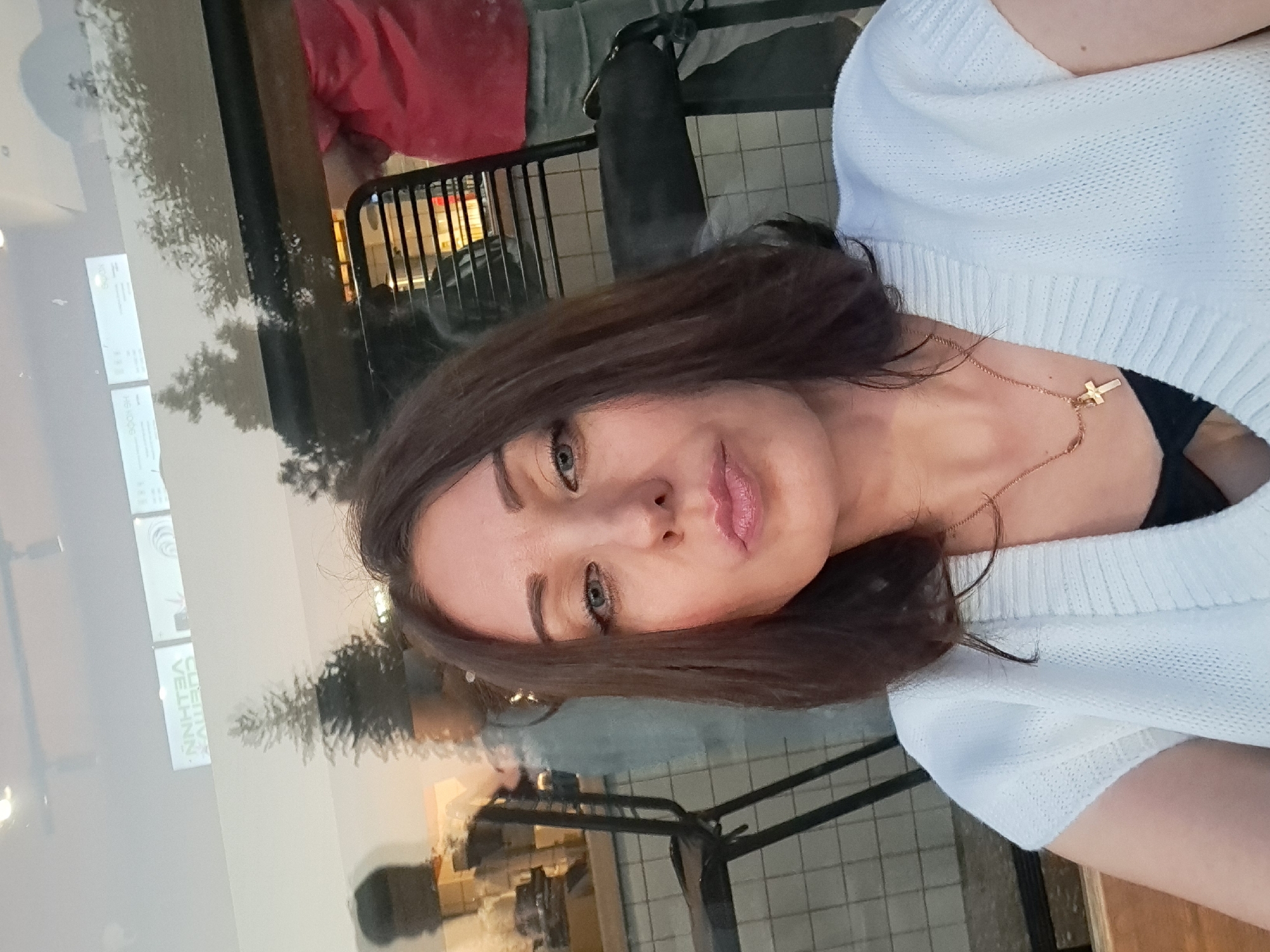 A woman with shoulder-length dark brown hair and blue eyes is taking a selfie indoors, possibly in a cafe or restaurant. She is wearing a white knitted top over a black garment and a gold cross necklace. Her lips are pursed, and she is looking directly at the camera. Reflections in the glass in front of her show trees, outdoor seating areas, and other people, including a person wearing a red shirt and another in a grey top. The background includes tiled walls and black chairs. The signs visible in the reflection appear to be in Cyrillic, suggesting the location is in Russia.