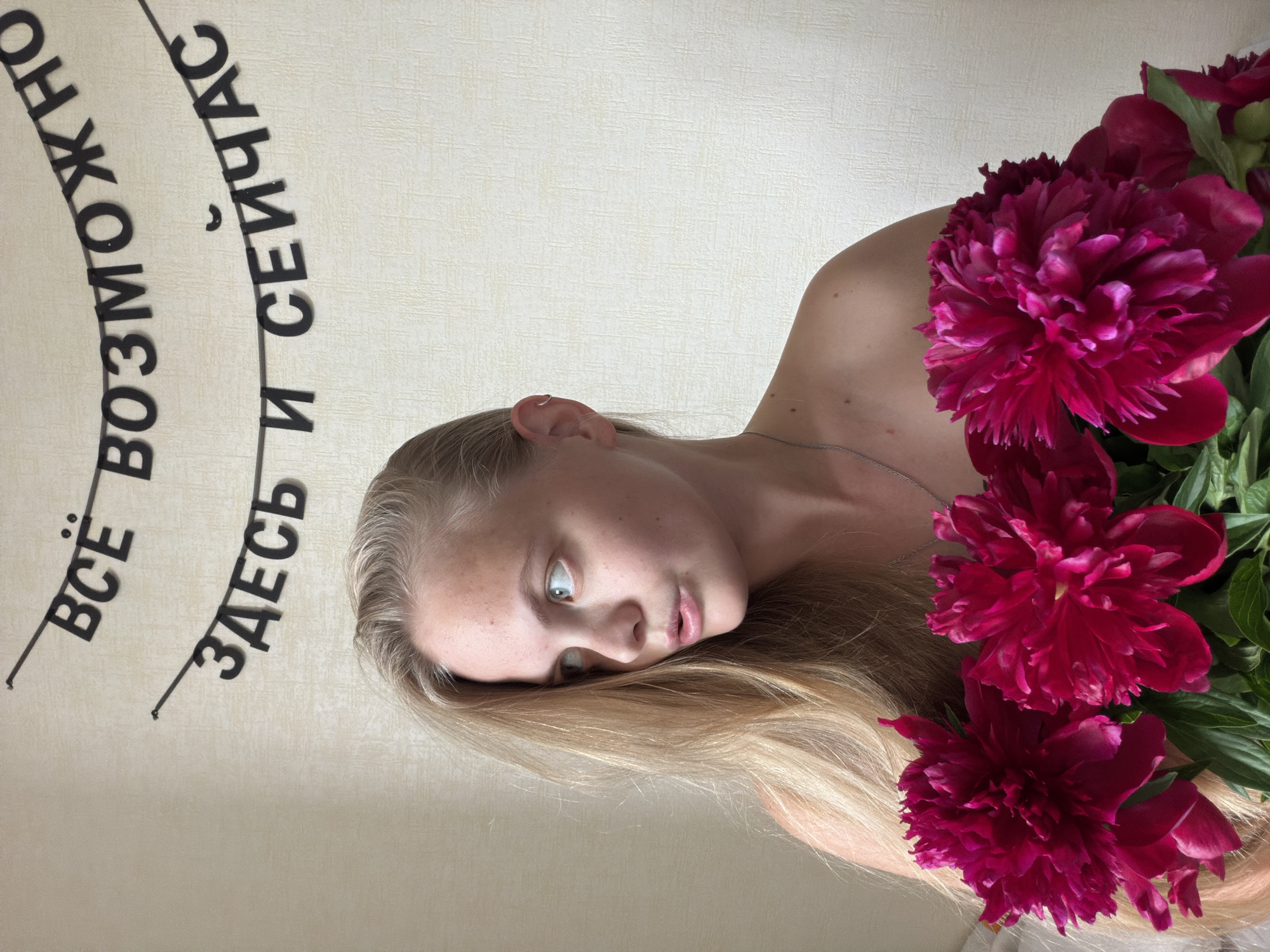 The original media file is oriented with a 90-degree clockwise rotation. When viewed upright, it features a close-up, artistic portrait of a young woman, likely in her late teens or early twenties, with long, light blonde hair. She appears to be lying down, looking directly upwards with a wide-eyed, perhaps surprised or contemplative expression. Her lips are slightly parted, and her fair skin shows subtle freckles. A delicate silver necklace is visible around her neck, and she is bare-shouldered.
To her right, partially covering her shoulder and hair, is a voluminous bouquet of deep red or magenta peonies. The flowers are vibrant, in full bloom, adding a rich splash of color and texture to the composition.
The background is a plain, light-colored (off-white or beige) wall with a subtle textured pattern, suggestive of wallpaper. On this wall, black, three-dimensional Cyrillic letters are arranged in an arc. The text reads "ВСЁ ВОЗМОЖНО ЗДЕСЬ И СЕЙЧАС" which translates to "Everything is possible here and now."
The lighting is soft and diffused, characteristic of an indoor setting. No specific time of day or weather conditions are discernible from the image. While the image itself lacks explicit geographical cues, it is contextually located in Izhevsk, Russia. The overall impression is a thoughtfully composed shot, possibly a selfie, conveying a sense of immediacy and potential, emphasized by the wall text.