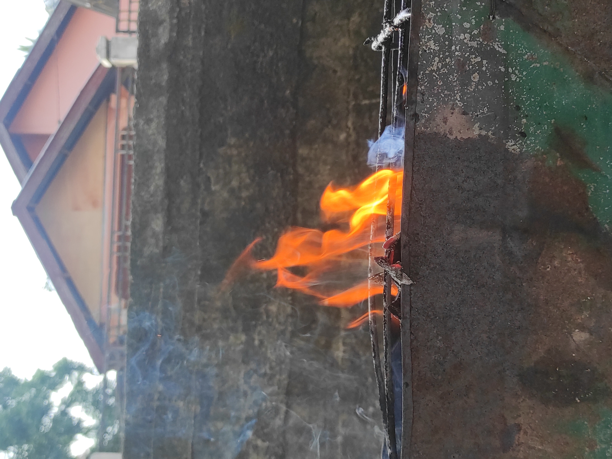 This vertical photograph, taken in Lhokseumawe, Indonesia, captures an outdoor scene dominated by a burning fire. In the foreground, the lower section of a weathered, dark gray-green metal container, likely a makeshift grill or pit, exhibits extensive rust and peeling paint. Above this container, a black metal grate supports several smoldering pieces of wood or charcoal, from which vivid orange flames rise, accompanied by distinct wisps of light blue-gray smoke originating primarily from the left side of the fire. Directly behind the fire and grill, a textured, dark gray concrete or stone wall extends horizontally across the midground. In the far background, above the wall, the peaked, dark reddish-brown roofs and light orange-peach walls of two residential buildings are visible against a bright, out-of-focus sky. Blurred green foliage is faintly discernible on the far left. No individuals are present in the frame.