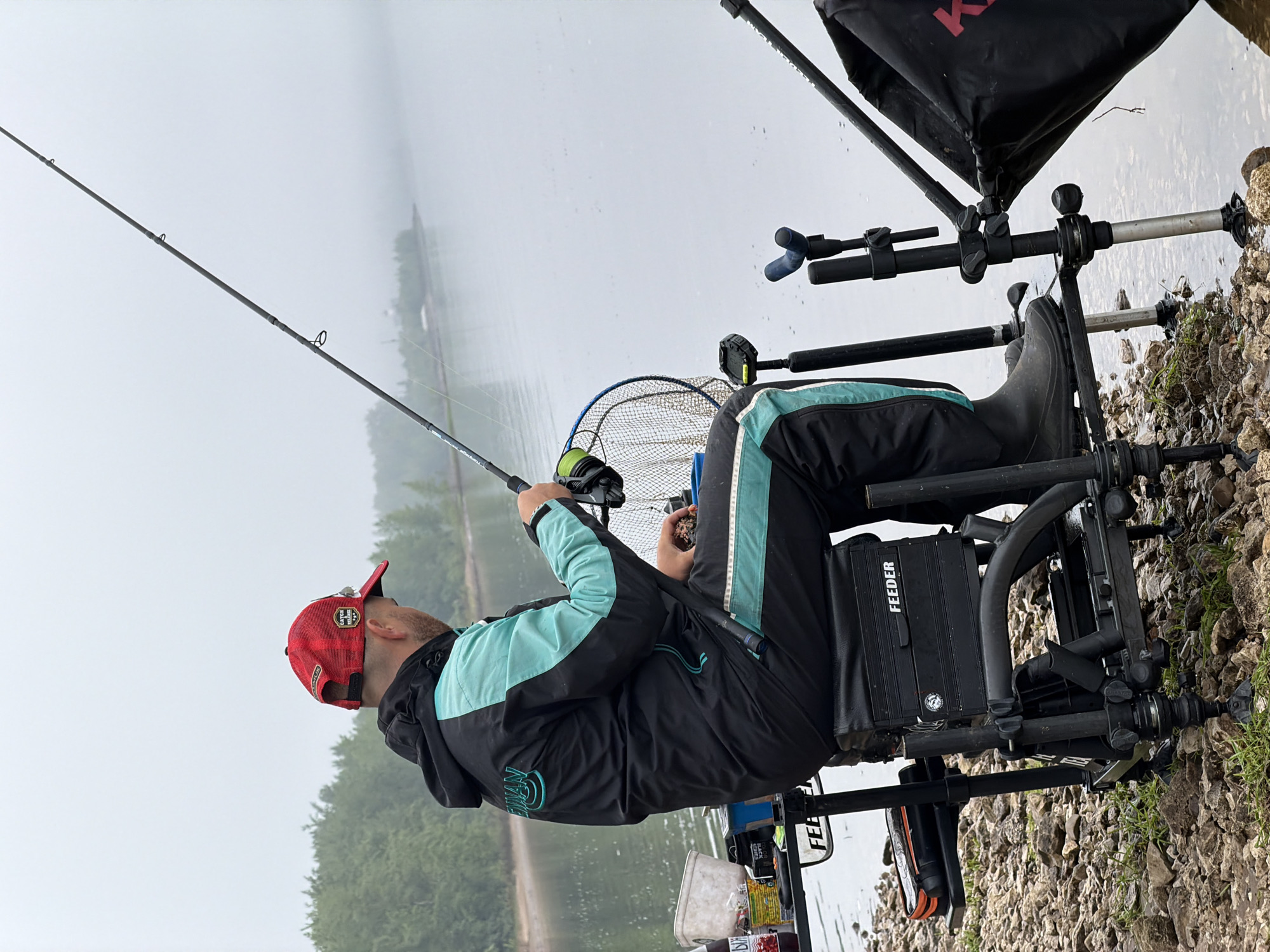 An angler is seated on a specialized fishing chair by the calm waters of a large lake or river. He is seen from the side, looking towards the water, holding a fishing rod with both hands. The man is wearing a black and turquoise fishing suit, a red baseball cap with a "SCOTCH & SODA" logo, and dark boots. A fishing reel is attached to the rod, and a green fishing net is visible behind him, near the chair.
The fishing chair is black and sturdy, equipped with various attachments, including a rod rest and a tackle box integrated into its design, with the word "FEEDER" visible on its side. Several containers and a white bucket with bait are placed around the chair. A large black bag, possibly a keepnet or tackle bag, is positioned next to the chair, with a partial "K" visible on it.
The setting is a rocky and pebbled shoreline, with the body of water extending into a hazy distance where distant trees are faintly visible on the opposite bank. The atmosphere appears misty or foggy, creating a soft, diffused light across the scene, suggesting either early morning or a day with low visibility. The man's jacket has a logo and the word "PRESTON" visible on it. He is actively engaged in fishing. The location context provided is Noviy, Russia.