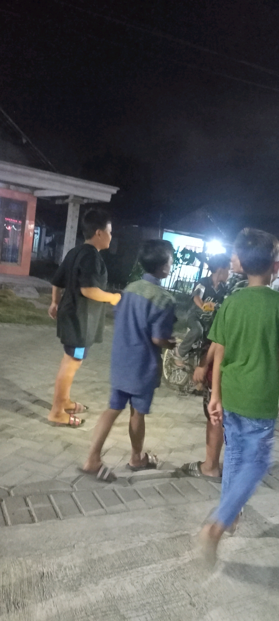 The image shows a group of four young boys standing on a paved area at night. The location is identified as the city of Gempolklutuk, Indonesia. The boys are dressed casually. The boy on the far left is wearing a black t-shirt and light-colored shorts with a blue accent. The boy next to him is wearing a blue shirt with a green patterned yoke and blue shorts. The boy on the far right is wearing a green t-shirt and blue jeans. Another boy, partially obscured, is wearing a black and white shirt and dark pants. There is a bicycle with a visible wheel in the middle ground, and a building with a light-colored roofline and a glass-fronted section is visible in the background. The ground is paved with rectangular interlocking stones. The lighting suggests it is evening or night, with artificial lights illuminating the scene.