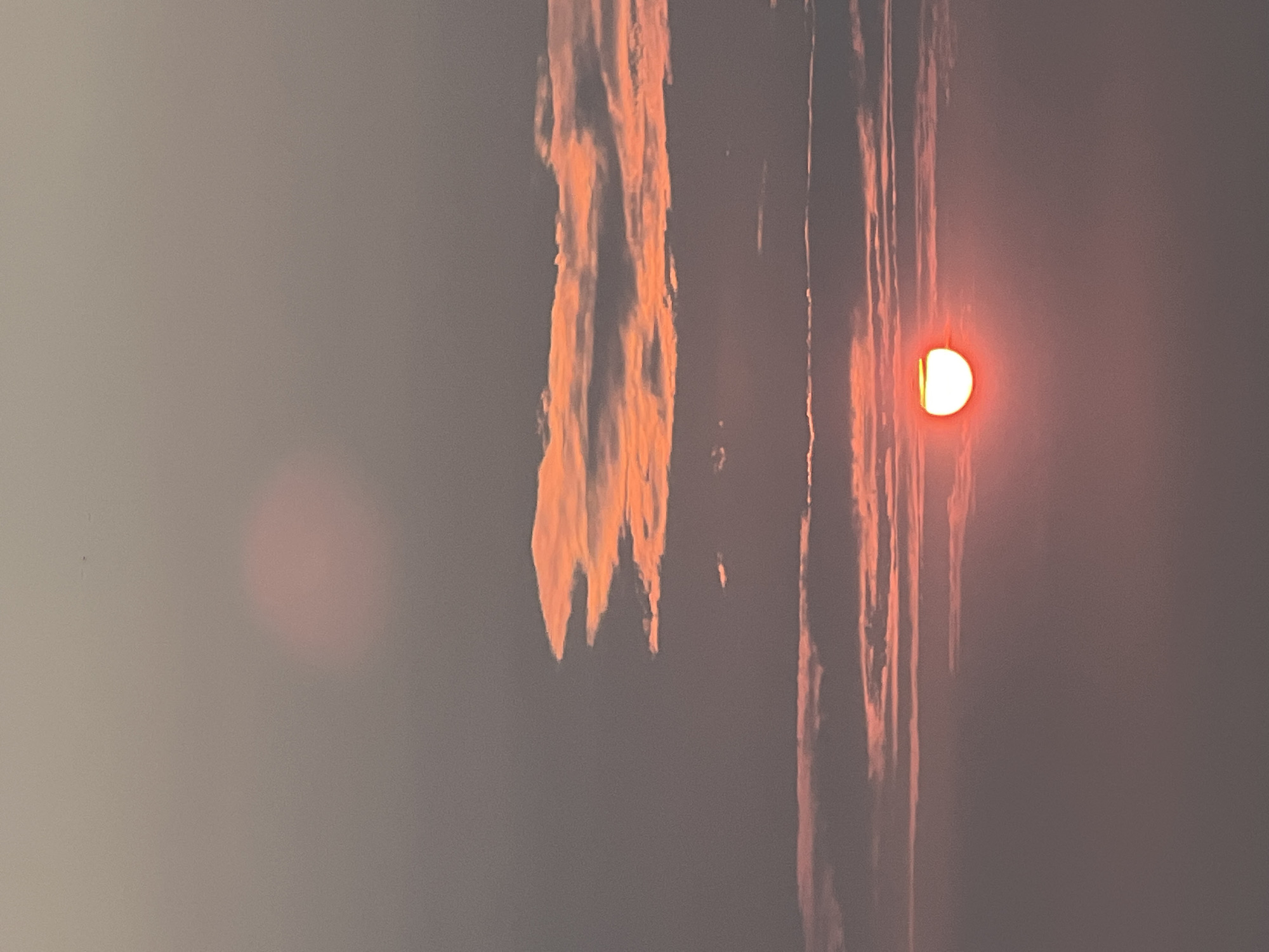 The image shows a sunset sky with several clouds. A bright orange sun is visible in the lower right quadrant of the image, partially obscured by clouds. The sky is a gradient of pink and gray, with some lighter pink areas indicating the direction of the sun. There are no people or man-made objects visible in the image. The scene appears to be an outdoor landscape.
