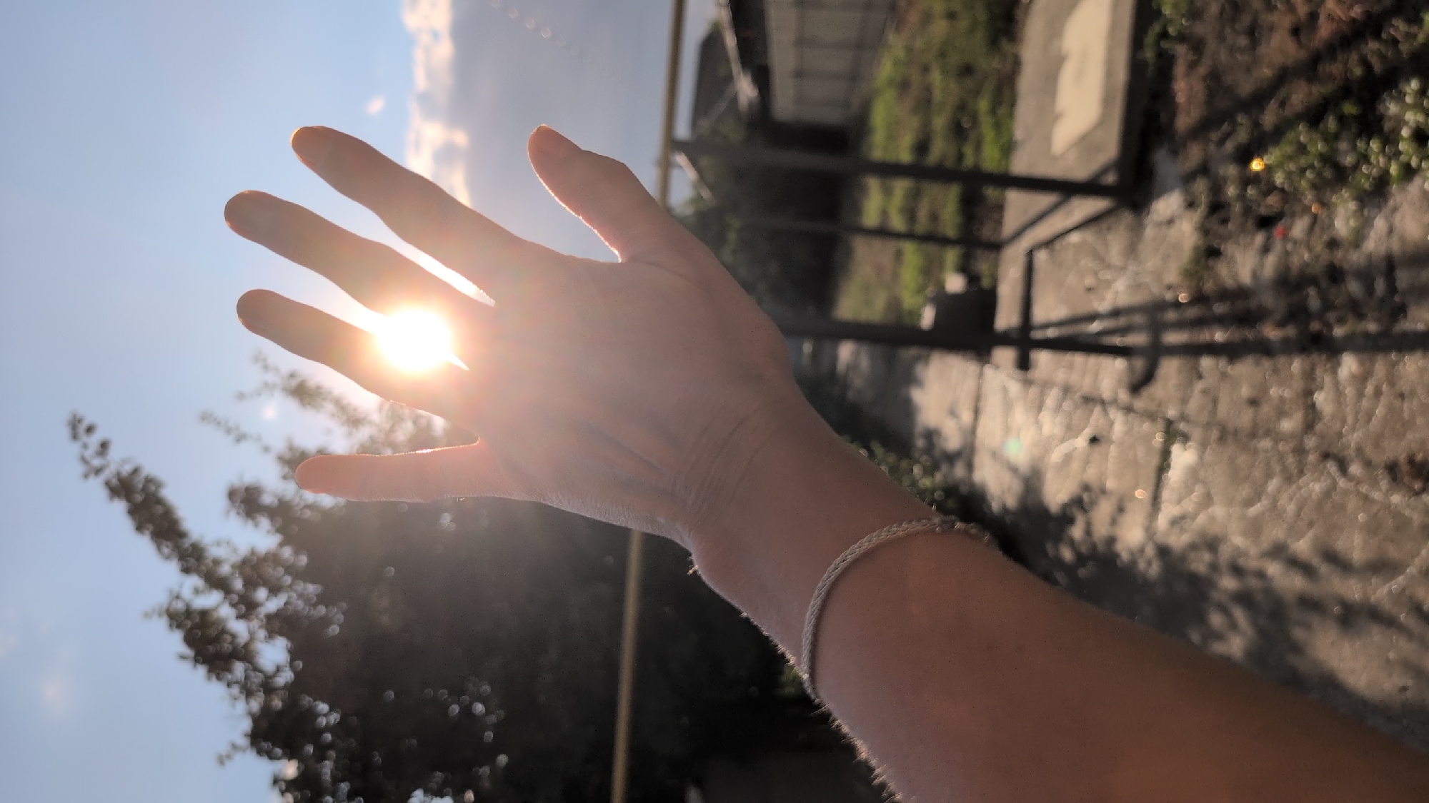The image is a close-up shot of a person's hand held up to the sun, blocking some of its rays. The sun is bright and positioned in the upper center of the frame, with visible lens flare. The hand is open, with fingers slightly spread, and a thin, braided bracelet is visible on the wrist. The background shows a blurred view of trees and a sky with clouds. The ground appears to be paved with concrete or stone. The overall lighting suggests it is daytime, likely late afternoon or early morning due to the angle of the sun and the warm tones. There is no specific information in the image that allows for the identification of the city of Izmail, Ukraine.