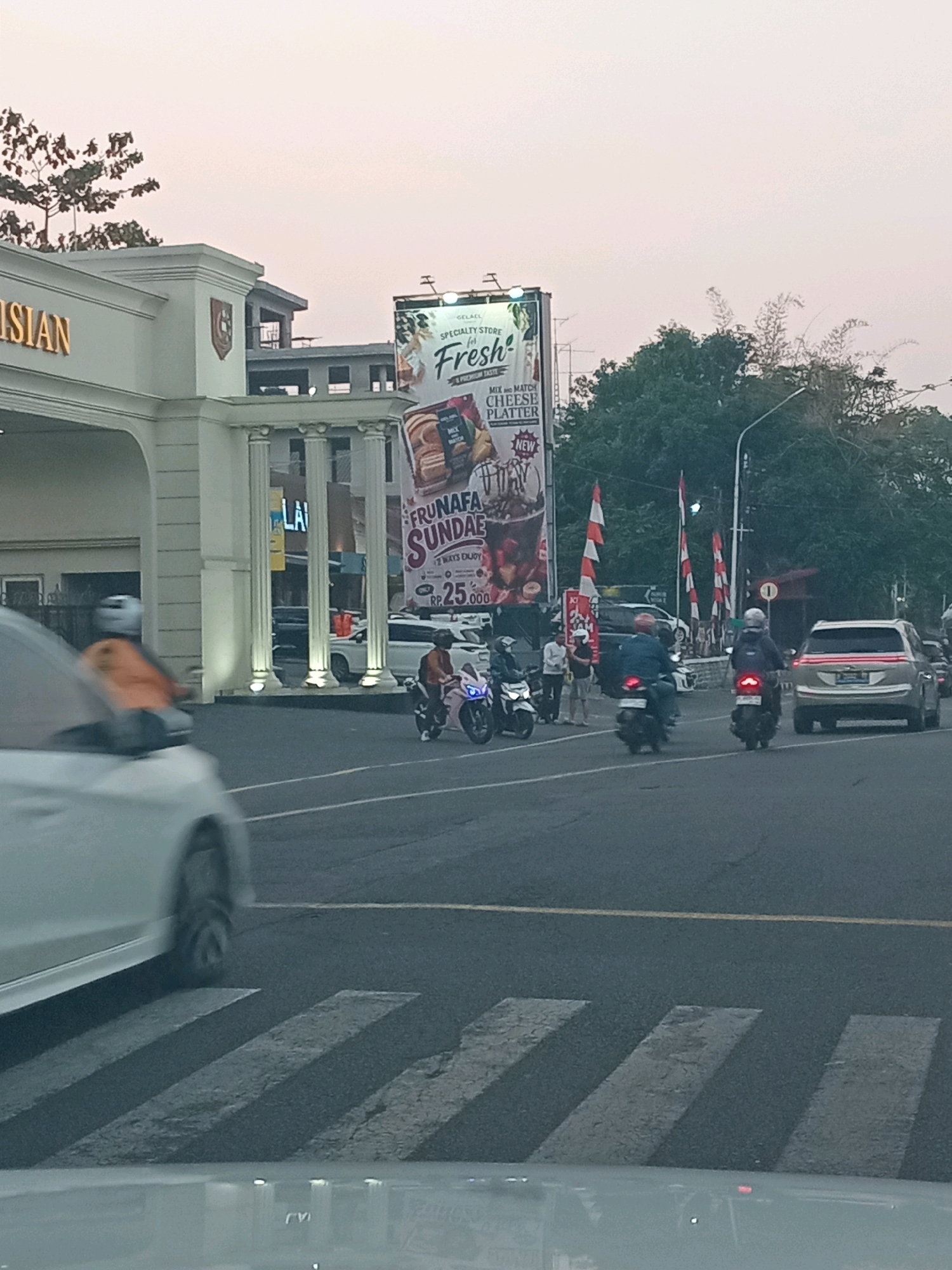 The image captures a street scene in Semarang, Indonesia, with a prominent white building featuring classical columns on the left. A large billboard for "Frunafa Sundae" is visible in the background, advertising a "Cheese Platter" for Rp 25,000. Several vehicles, including motorcycles and cars, are on the road. Two people are riding motorcycles, and a few individuals are standing near the billboard. The sky is overcast, suggesting it might be dawn or dusk. The foreground shows a car's windshield and a crosswalk.