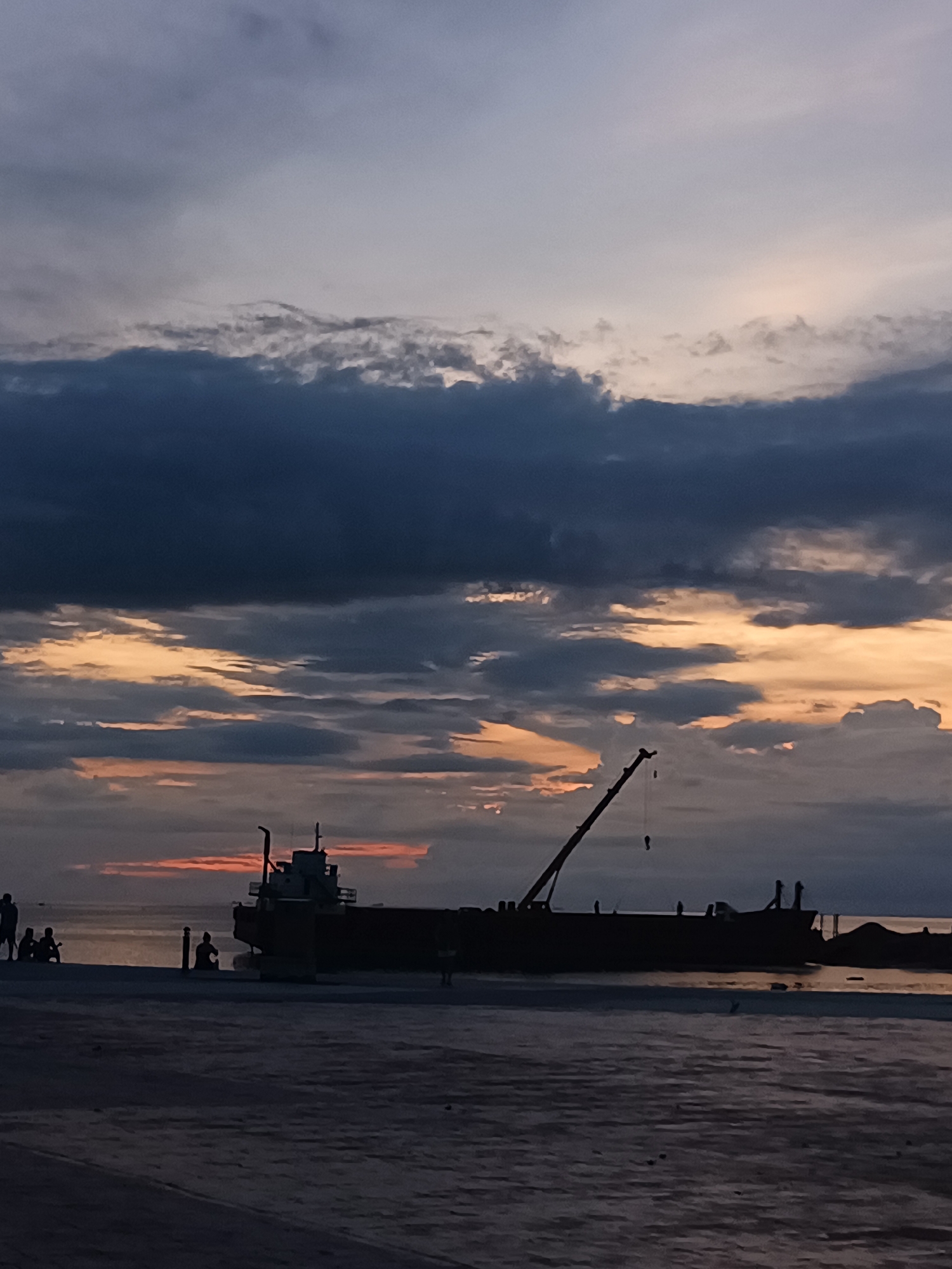 The image depicts a dusk scene at what appears to be a harbor or pier in Koh Phangan, Thailand. Silhouetted against a dramatic sky with streaks of orange and yellow peeking through dark clouds, a large barge with an extended crane dominates the mid-ground. Several figures are visible on the foreground pier, which is paved with a textured surface. To the left of the barge, a small group of people are gathered, appearing to be observing the water or the barge. To the right of the barge, a single person is seated on the pier. The water reflects the muted colors of the sky. The overall atmosphere is tranquil and serene, characteristic of a sunset at a coastal location.
