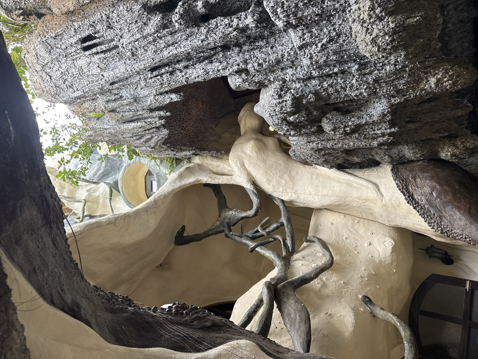 The image shows the exterior of the Crazy House in Da Lat, Vietnam. The architecture is organic and surreal, resembling a cluster of caves and trees. The foreground features a large, dark, textured structure on the right that appears to be made of concrete or a similar material, designed to mimic rough rock or bark. Overhanging this is a dark, textured archway, also likely constructed material. To the left, smooth, cream-colored walls curve and flow, mimicking the shapes of natural formations. Intertwined within these structures are dark, gnarled branches, suggesting a fusion of natural and man-made elements. Sunlight filters down from above, highlighting the textures and forms. In the background, glimpses of rounded windows with dark frames are visible within the cream-colored architecture, and some green foliage sprouts from the top. The overall impression is one of an otherworldly, organic structure, integrated with its natural surroundings.