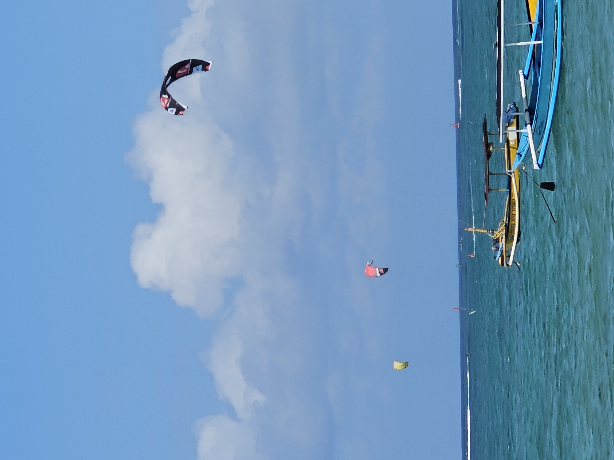 This image captures a vibrant scene at a beach in Denpasar, Indonesia, under a bright blue sky dotted with fluffy white clouds. The turquoise water of the ocean stretches out, with gentle waves lapping at the shore. Several traditional Indonesian boats, known as jukung, are moored or gently bobbing on the water, some painted in bright yellow and blue. One jukung, in the foreground and painted blue, has a white railing and an oar resting inside. Behind it, a yellow jukung has a thatched roof and appears to be labeled "KAISER SURF SCHOOL".
The sky is alive with activity as multiple kite surfers are seen in the distance. Three large, colorful kites are visible: one predominantly black and red, another vibrant red, and a third a bright yellow. These kites are tethered to surfers who are either riding the waves or preparing to do so, their lines connecting them to the kites. In the far distance, a white boat creates a wake as it moves across the water, and a small red buoy is visible near the horizon. The image conveys a sense of recreation and a beautiful day at the beach in Denpasar.
