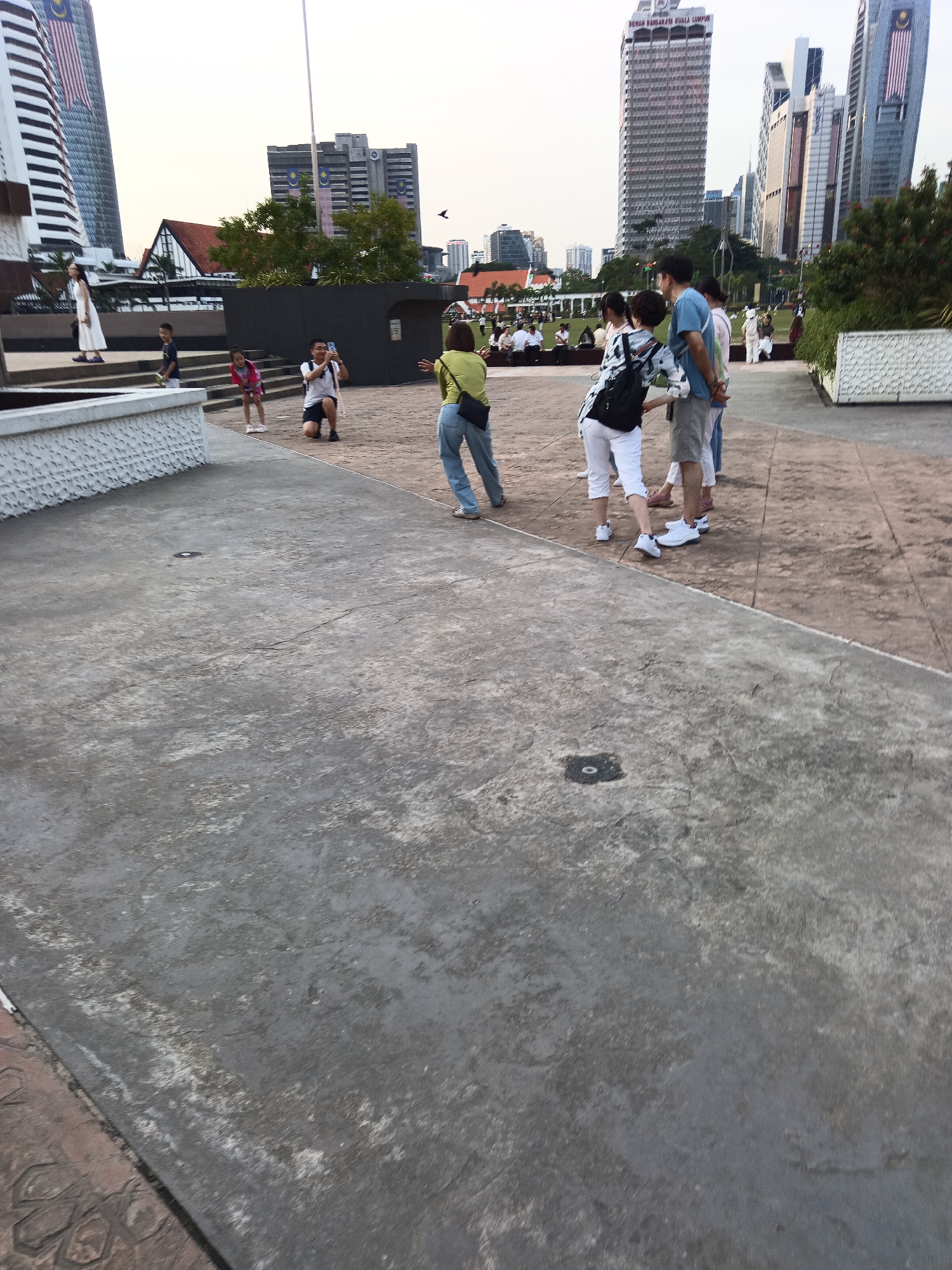 The image depicts a scene in Kuala Lumpur, Malaysia, likely at Independence Square (Dataran Merdeka) due to the presence of high-rise buildings with Malaysian flags and the general urban park setting. In the foreground, a grey concrete pathway dominates the lower half of the frame. To the right, a reddish-brown paved area is visible.
Several people are present. On the left, a woman in a white dress stands near some stairs. Two children are positioned in front of her, one in a blue shirt and another in a pink top. A man is kneeling on the reddish-brown pavement, holding a phone as if taking a photograph. Further to the right, a group of people are standing and walking. A woman in a lime green top and blue jeans is facing away from the camera, as is a man in a blue t-shirt and grey shorts. Another woman in a white and black top and white pants is also with this group.
In the background, several modern skyscrapers are visible against a pale sky. Prominent among them is a tall, white building with a checkered facade and a Malaysian flag. Another distinctive building with a cylindrical design and Malaysian flags is also visible. Trees and other buildings can be seen throughout the cityscape. The overall impression is an outdoor public space with people engaging in leisure activities and sightseeing in an urban environment.