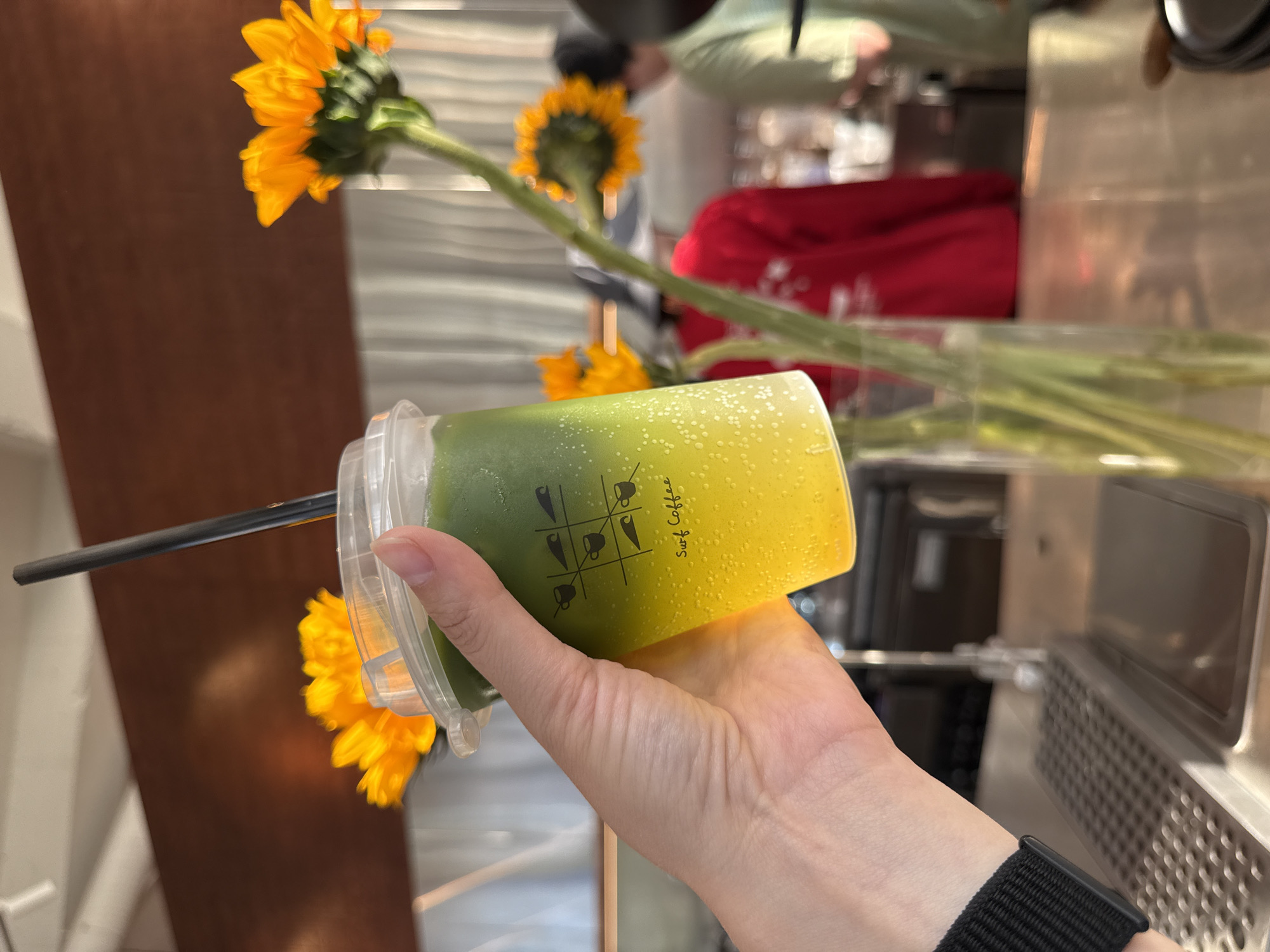 A hand holds a plastic cup of a layered green and yellow beverage with a black straw in front of a vase of sunflowers. The cup has "Surf Coffee" printed on the side. The background appears to be a cafe interior, including metal surfaces and a red object. The image was taken at a location within the city of Кощаково, Russia.