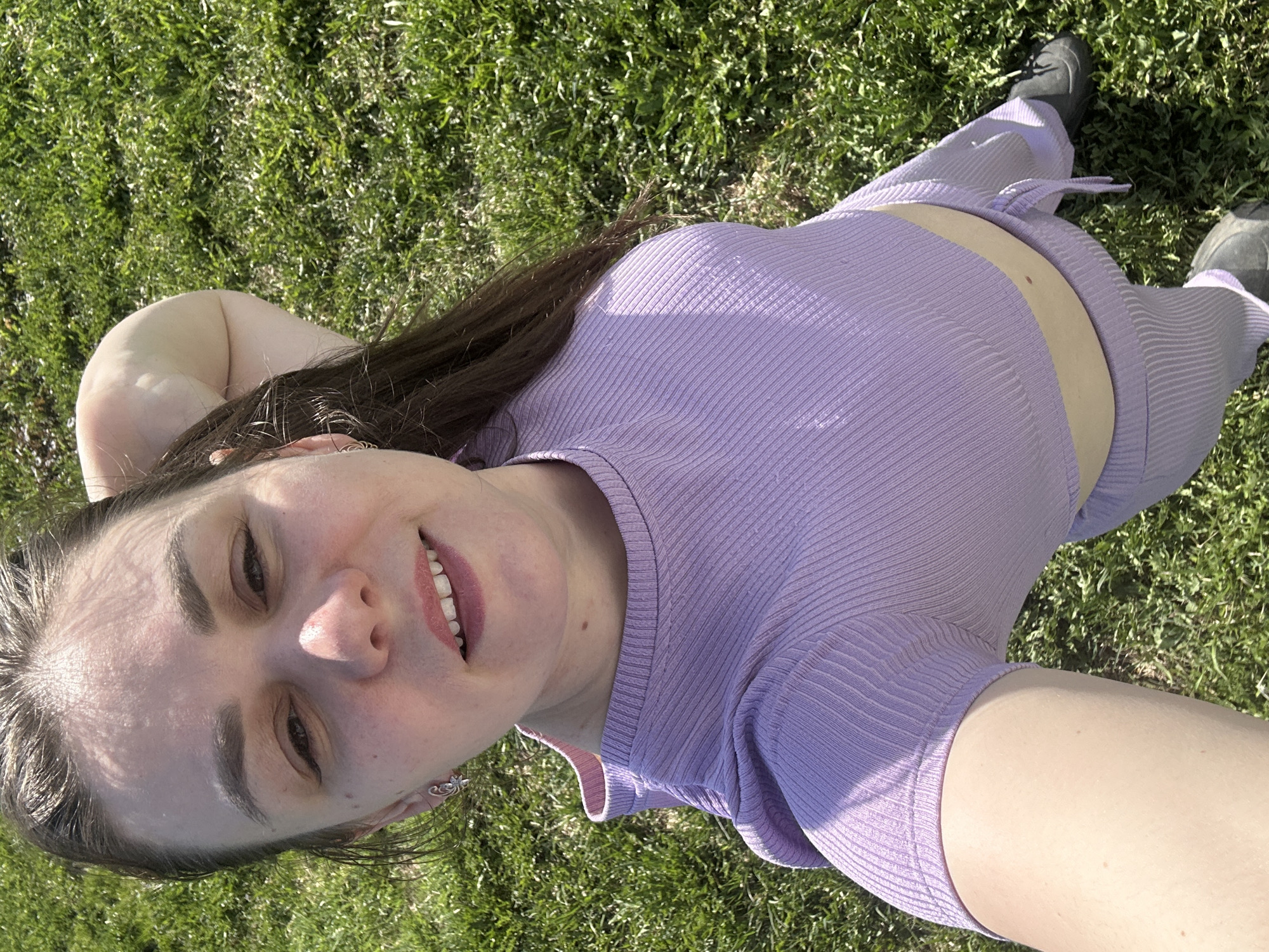 A woman is lying on a grassy area in Chaikovsky, Russia. She is wearing a light purple ribbed two-piece outfit consisting of a short-sleeved top and pants. She has dark hair and is smiling at the camera. She is wearing sneakers. The background is green grass.