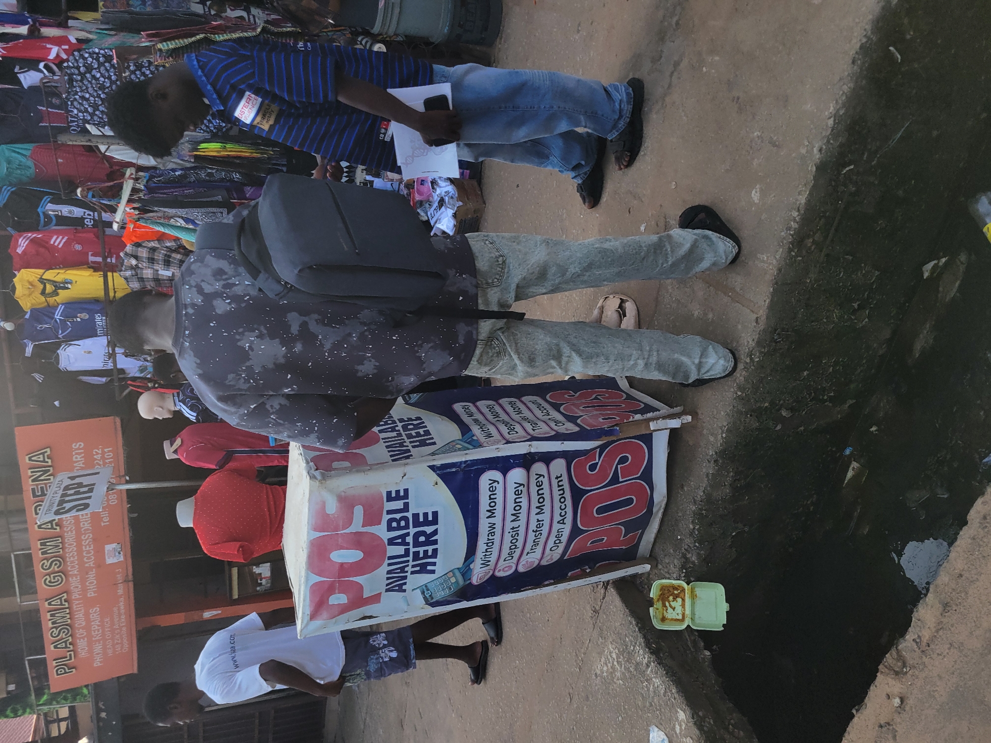 The image depicts an outdoor street scene in Egbagu, Nigeria, with a focus on a POS (Point of Sale) service stand. Three men are present. One man, wearing a white t-shirt with "www.sea.com" visible and patterned shorts, stands to the left, looking towards the POS stand. Another man, wearing a grey splatter-patterned t-shirt and light-colored jeans, stands behind the POS stand, holding a dark backpack. The third man, wearing a blue and black striped t-shirt and blue jeans, stands to the right of the POS stand, holding a document and a dark phone.
The POS stand is a tall, cylindrical object wrapped in a blue and white advertisement promoting POS services. Visible text on the stand includes "POS AVAILABLE HERE" and lists services such as "Withdraw Money," "Deposit Money," "Transfer Money," and "Open Account."
In the background, a shop with an orange sign that reads "PLASMA GSM ARENA (HOME OF QUALITY PHONE ACCESSORIES/SPARE PARTS, PHONE REPAIRS, PHONE ACCESSORIES DRIVE...)" is visible. Various clothing items are displayed for sale on racks in front of this shop. A drainage ditch is present in the foreground, containing murky water and some debris. A light green food container with some contents is placed on the edge of the ditch. The ground is a mixture of concrete and dirt.