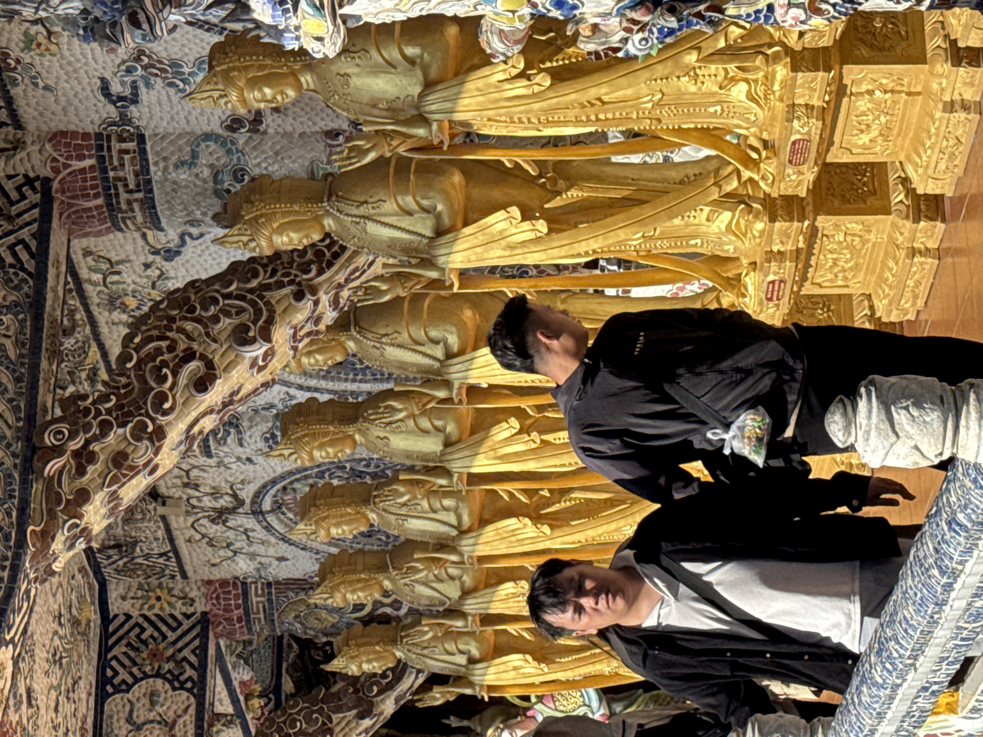 The image depicts a row of golden statues of Buddhist deities, likely bodhisattvas, standing in a temple or ornate shrine. The statues are adorned with elaborate headdresses and jewelry, and their robes flow dramatically. The background wall is intricately decorated with colorful mosaic tiles featuring floral and geometric patterns, as well as dragon motifs. The architectural elements of the shrine, including carved wooden beams and tiled ceilings, are also highly decorative.
In the foreground, two men are visible. The man on the left, facing the camera, wears a dark jacket over a light-colored collared shirt. He has short, dark hair and a neutral expression. The man on the right, facing away from the camera and looking towards the statues, wears a dark jacket and black pants. He has short, dark hair and is carrying a small, clear plastic bag with colorful contents slung over his shoulder. Both men are standing on a tiled walkway behind a decorative blue and white tiled railing. The overall scene suggests a place of cultural and religious significance in Xuân Thành, Vietnam.