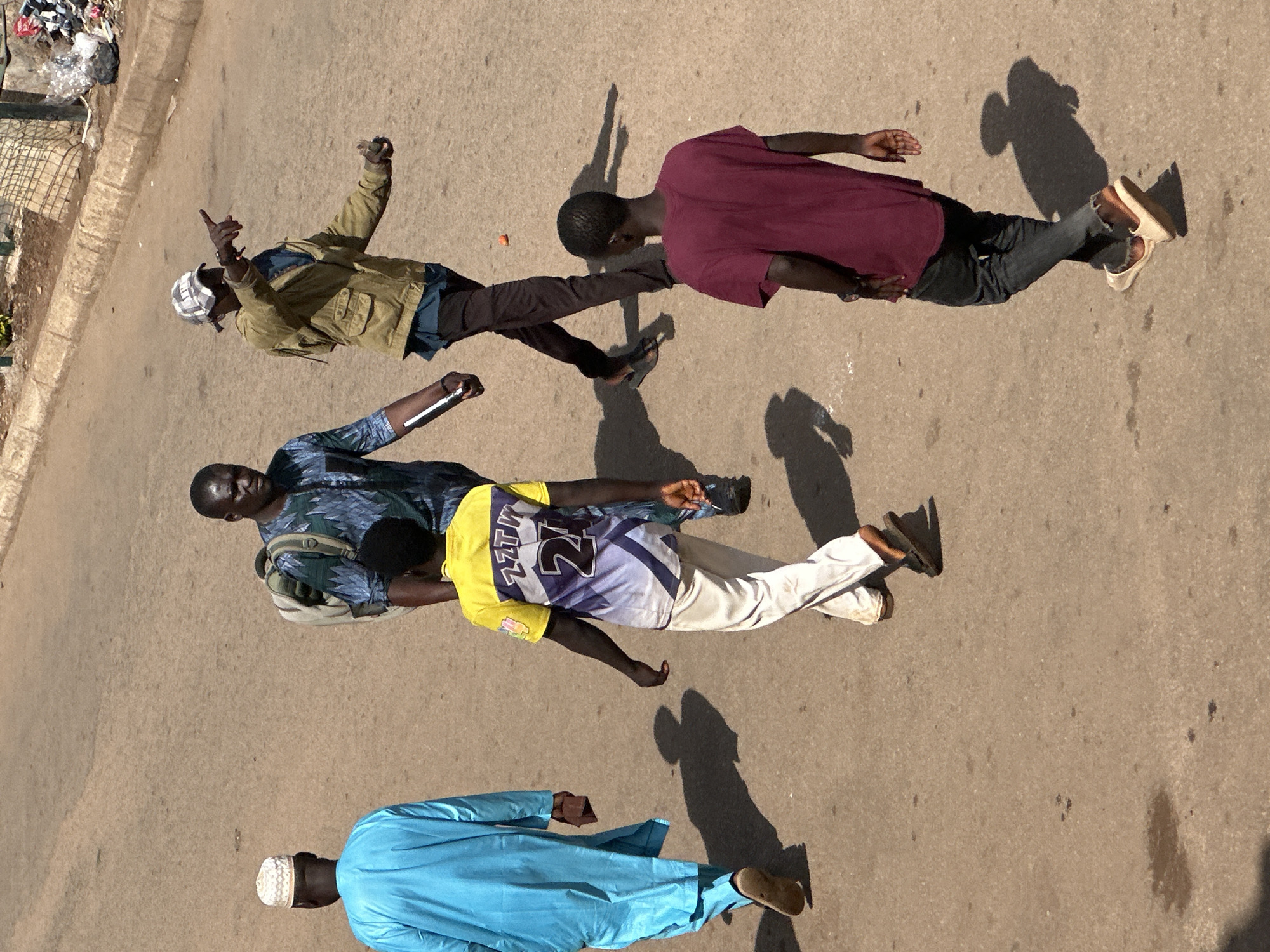 A group of five men are walking on a paved street in Bukuru, Nigeria. The man on the far left is wearing a blue robe and a white cap. Next to him, a man in a blue and green patterned shirt walks with a backpack. A third man, in a yellow and purple shirt with the number 24, walks ahead. Further ahead, a man in a tan jacket points to his right. The man closest to the camera wears a burgundy t-shirt and dark jeans. The environment appears to be an urban street with a sidewalk on the right side, featuring some litter. The lighting suggests it is daytime.
