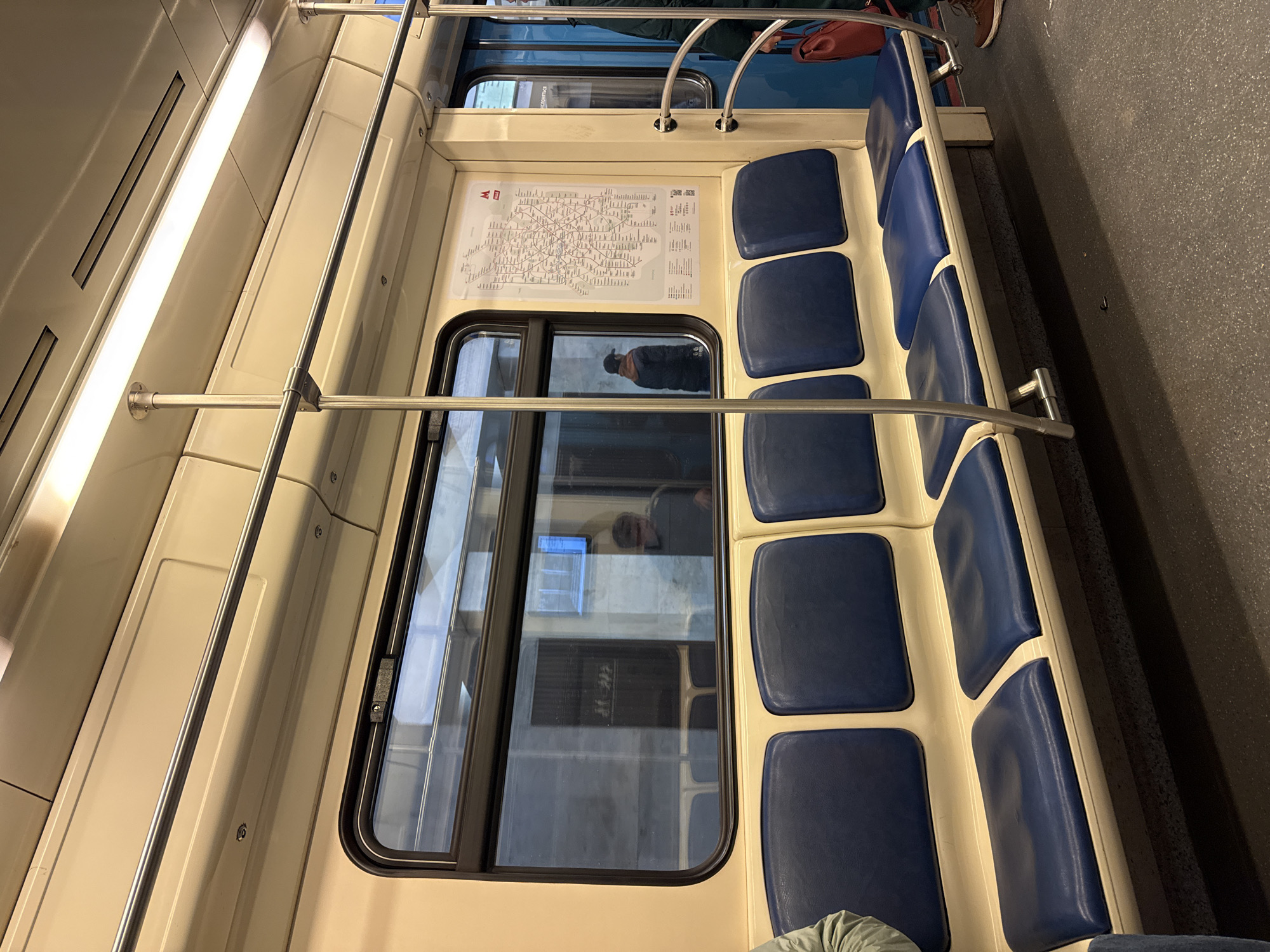 The image displays the interior of a subway car, specifically identified as part of the Moscow Metro system by a prominent map on the wall. The car features a row of blue-cushioned seats with cream-colored frames, extending along one side. Above the seats, and throughout the ceiling, are several silver handrails and poles for passengers to hold. The ceiling itself is cream-colored and illuminated by long, bright fluorescent light strips. The floor is dark gray with a textured appearance.
A large window dominates the central wall area, offering reflections of the train's interior. In these reflections, at least two individuals are visible; one person, wearing a dark cap, is clearly reflected, while another is more indistinct. Outside the window, faint architectural elements can be glimpsed, suggesting it is daytime.
A detailed map of the Moscow Metro network is affixed to the wall above the window. The map prominently displays the red "М" logo of the Moscow Metro in its top-left corner. Numerous station names and line diagrams, written in Cyrillic script, cover the map. Adjacent to the map, several QR codes and additional small text, also in Cyrillic, are visible.
Other partial subjects include a person's foot in a brown shoe and a red bag at the top right, and a person wearing a green garment at the bottom left. The scene depicts a functional public transport environment.