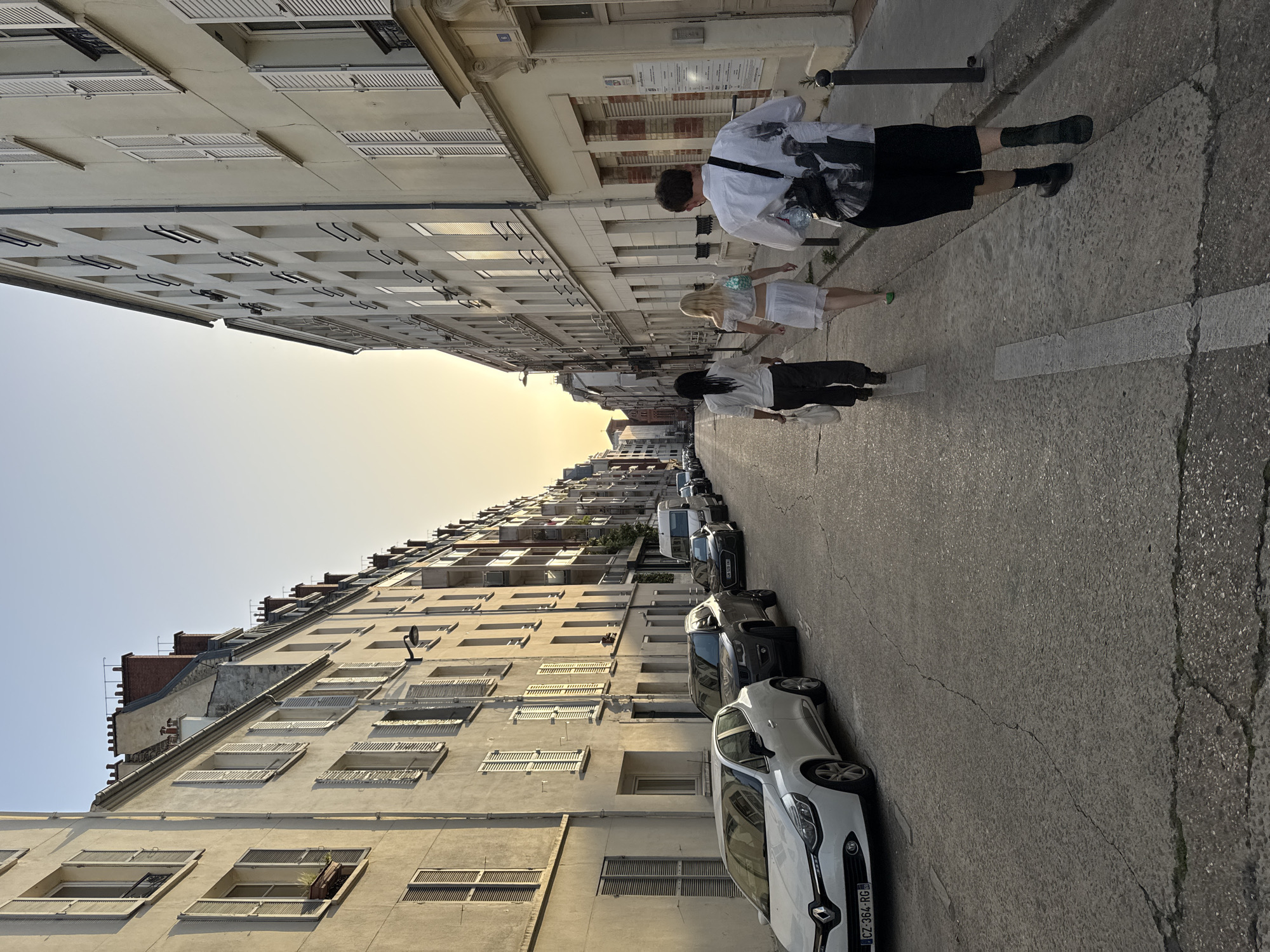 A street scene in Paris, France is captured with buildings lining both sides of a road. Cars are parked on the right side of the street. Three people are walking in the opposite direction on the right-hand side of the road, away from the camera. The first person is wearing a white shirt and black shorts, carrying a bag over their shoulder. The second person is wearing a white dress, and the third person is wearing a white shirt and black skirt, carrying a white bag. The sky is visible in the distance and appears to be during golden hour.