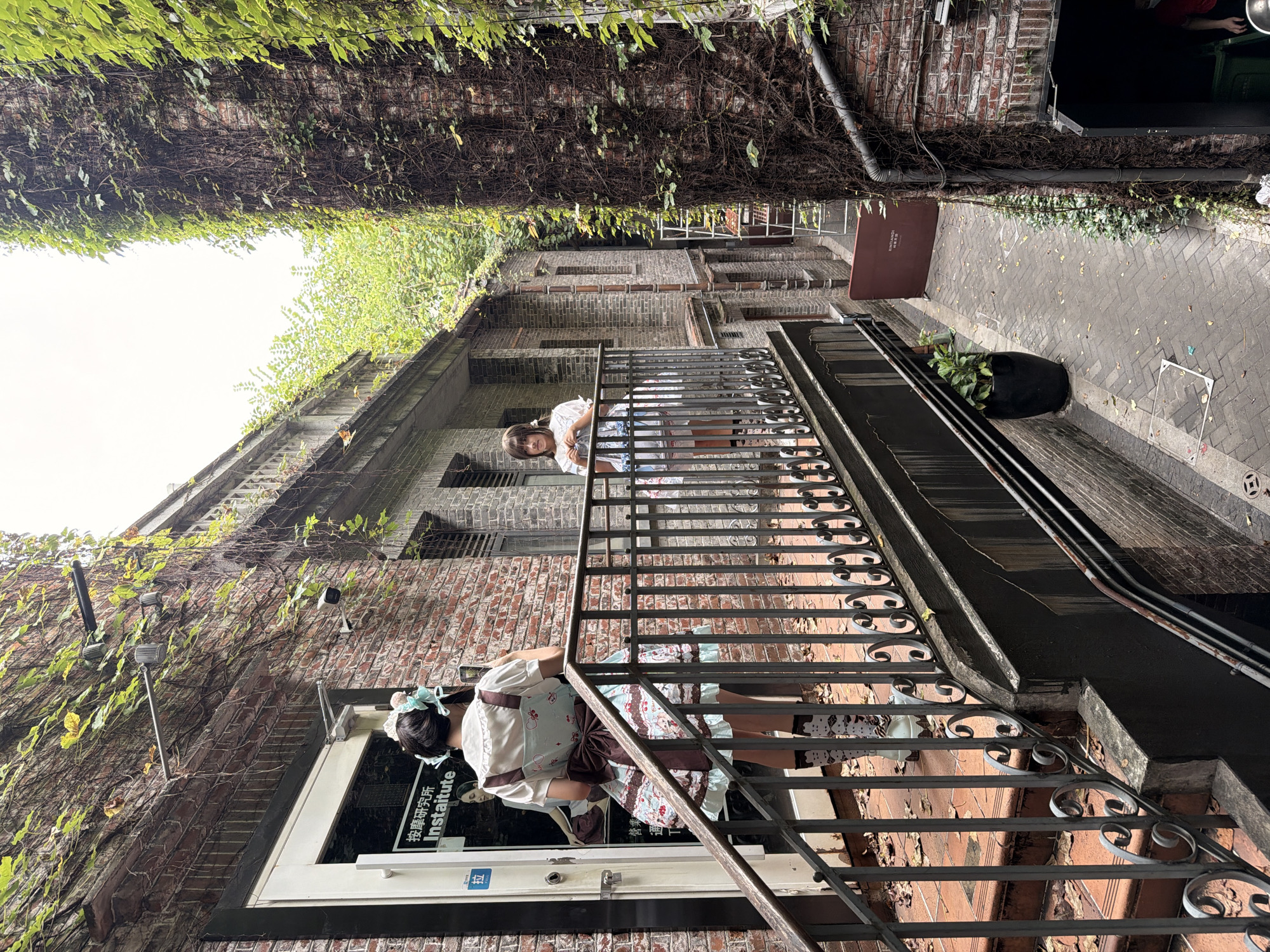 The image depicts a narrow alleyway in Chancheng District, China, flanked by brick buildings covered in lush green ivy. In the foreground, a young woman in a Lolita-style dress, complete with a large bow and patterned stockings, is captured from behind as she ascends a metal-railed staircase. She is holding a smartphone, seemingly taking a photo. To her right, across the alley and on a balcony, another young woman in a white dress and with dark hair stands, holding a blue bag. The alleyway itself is paved with bricks and scattered with fallen leaves. A large black planter with a green plant sits near the bottom left of the frame. The overall atmosphere is one of an urban, somewhat overgrown, yet charming historical district.