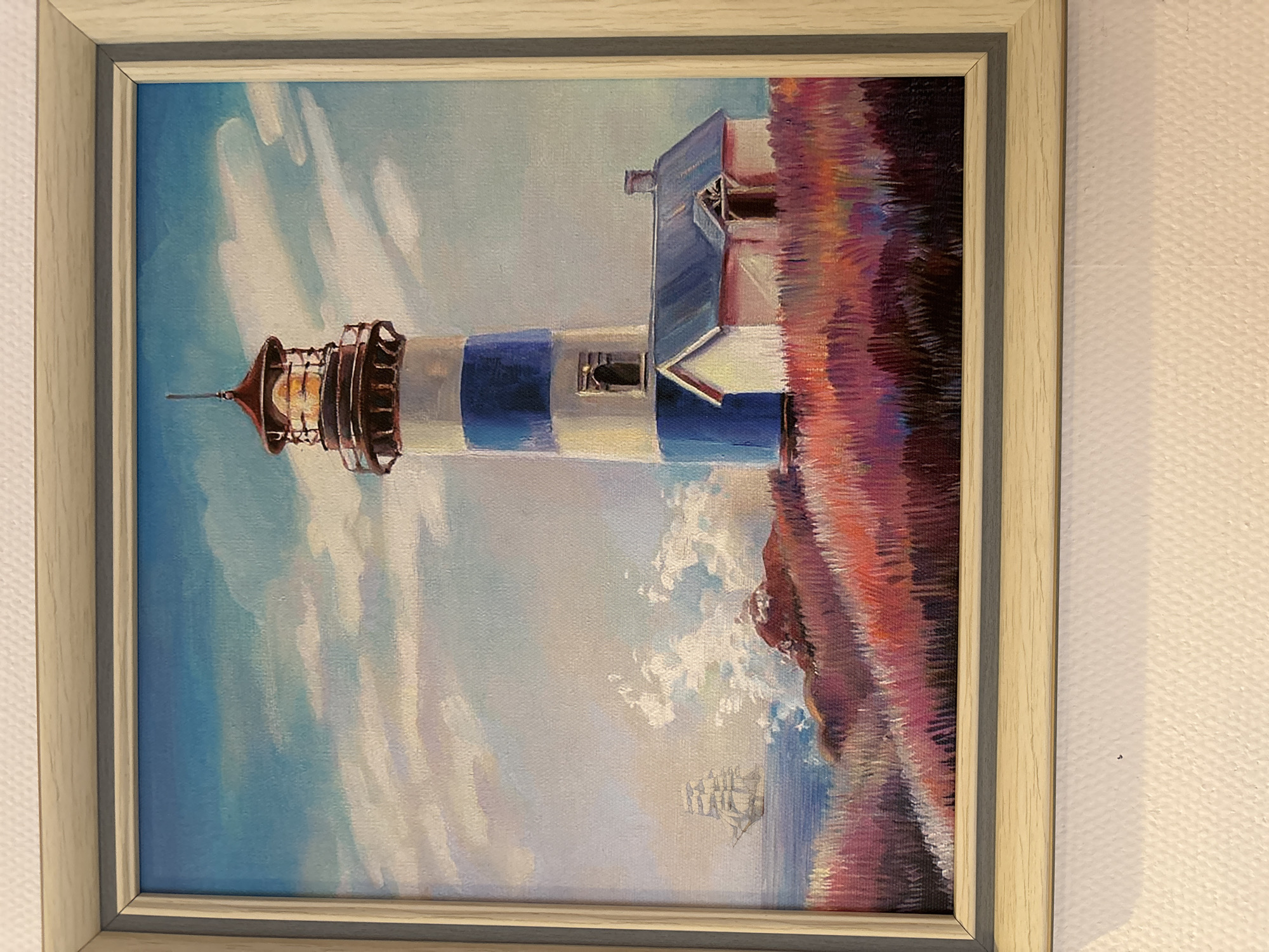 A framed painting, depicting a vibrant maritime landscape, is mounted on a textured, off-white wall. The painting's central subject is a tall, cylindrical lighthouse, distinguished by its white body and two prominent blue horizontal stripes. The lighthouse's lantern room, rendered in dark brown, emits an internal orange glow. Adjacent to the lighthouse base is a small, white single-story house with a blue sloped roof and a dark chimney. Both structures are positioned on a hilly foreground covered in dense, brush-stroked vegetation, featuring hues of red, pink, purple, and orange. To the left of the lighthouse, the sea is visible with white-capped waves crashing against a rocky coastline. Further in the distance on the water, a multi-masted sailing ship is depicted. The sky above is a light blue, sparsely populated with elongated white clouds. The painting is enclosed by a simple, light-colored, possibly wooden, rectangular frame. This framed painting is situated on a wall within the city of Русский Вожой, Russia.