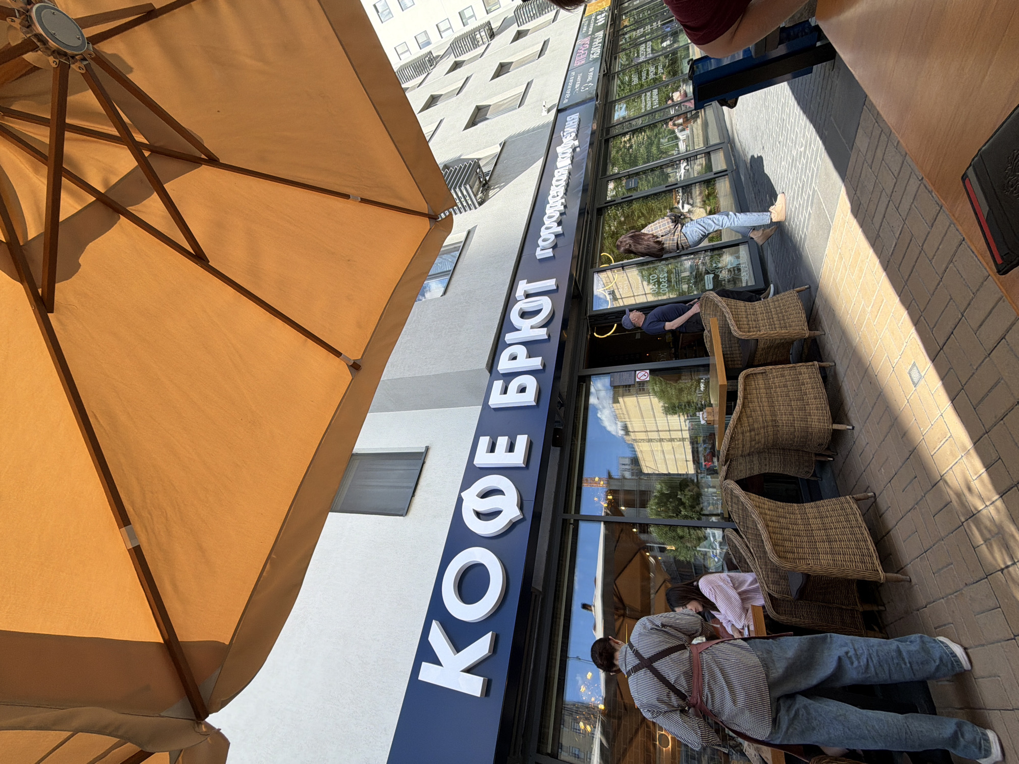 This image captures an outdoor scene in what appears to be a bustling city street, specifically Tyumen, Russia, during bright daylight. The primary focus is an outdoor seating area of a cafe or coffee shop.
In the immediate foreground, a large, light brown or beige patio umbrella provides shade, with its internal wooden structure visible. Part of a wooden table is also seen, suggesting a person is seated beneath the umbrella, with only a maroon sleeve visible.
The midground features the facade of a building with a prominent dark blue sign displaying white Cyrillic letters. The main text reads "КОФЕ БРЮТ" (KOFE BRYUT), which translates to "Coffee Brut." Above this, smaller Cyrillic text is visible, reading "ГОРОДСКОЙ ПАРАДОКС" (GORODSKOY PARADOKS), meaning "City Paradox." The cafe entrance features large glass windows that reflect the surrounding buildings and trees.
Several individuals are present. One person in a checkered shirt and blue jeans, wearing an apron-like harness, stands near the cafe entrance, possibly staff attending to customers or waiting. Another person in a dark shirt and cap is seated on one of the outdoor wicker chairs, looking towards the cafe. Further down, another person in a plaid shirt and jeans is seen walking past the cafe entrance. The outdoor seating consists of multiple wicker chairs and small tables arranged on a paved sidewalk made of light-colored bricks.
The background shows a multi-story building with numerous windows, indicating an urban setting. Strong shadows are cast across the sidewalk and chairs, suggesting a sunny day, likely in the morning or late afternoon. The overall atmosphere is that of a relaxed urban environment, with people enjoying the outdoor space.