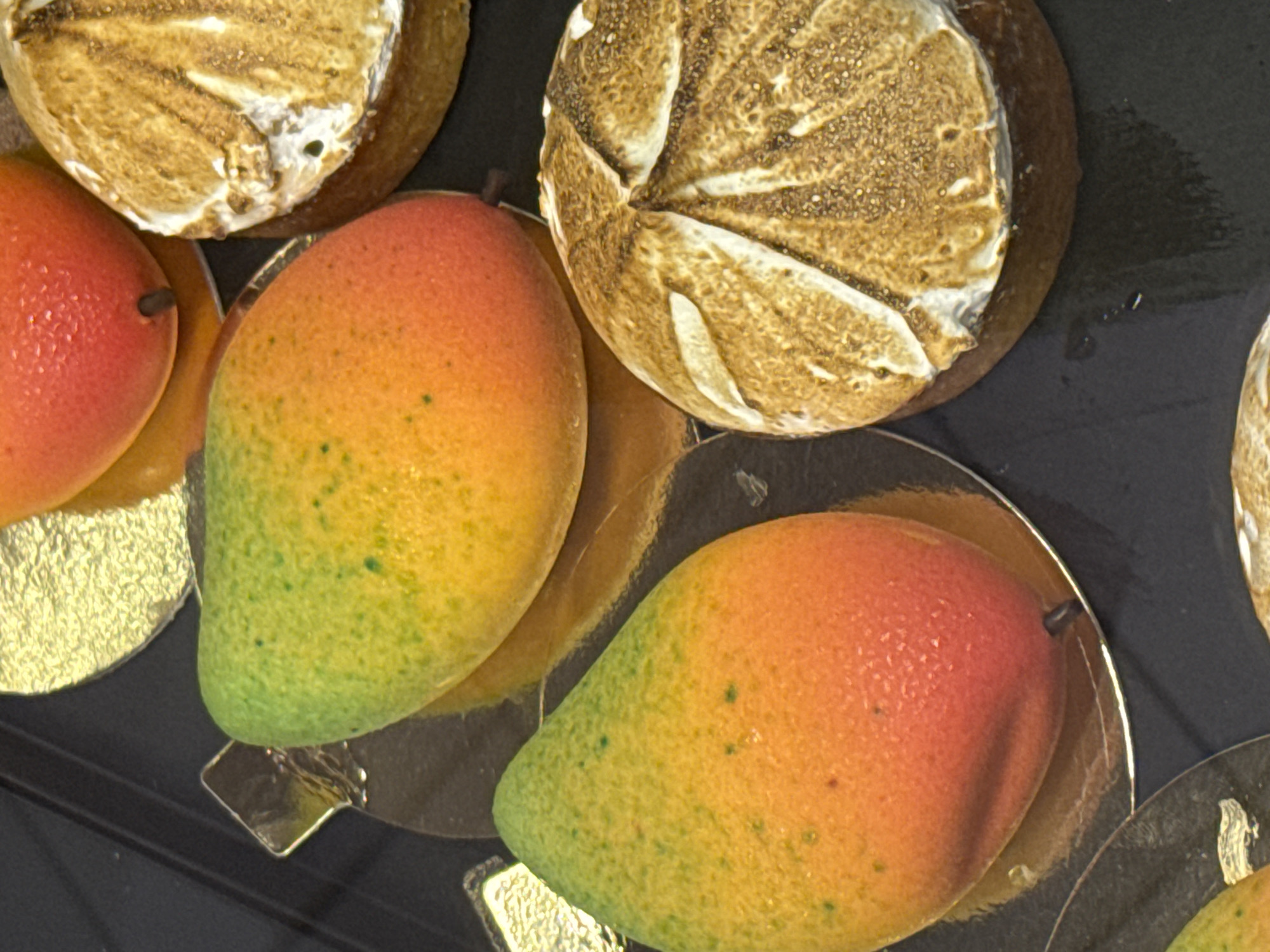 The image shows a close-up of several artisanal desserts, likely in a display case. Two prominent desserts are shaped like mangoes with a gradient of green, yellow, and red colors, sitting on gold-colored circular bases. To the right, there are two round desserts topped with toasted meringue, creating a textured, golden-brown surface with white swirls. The desserts are arranged on a dark, reflective surface, possibly a countertop or a display shelf. There are no people visible in the image. The scene appears to be a detailed shot of food presentation in a culinary establishment, possibly a bakery or a patisserie. Without further context or external information, it is impossible to definitively determine the specific location in Budva, Montenegro.