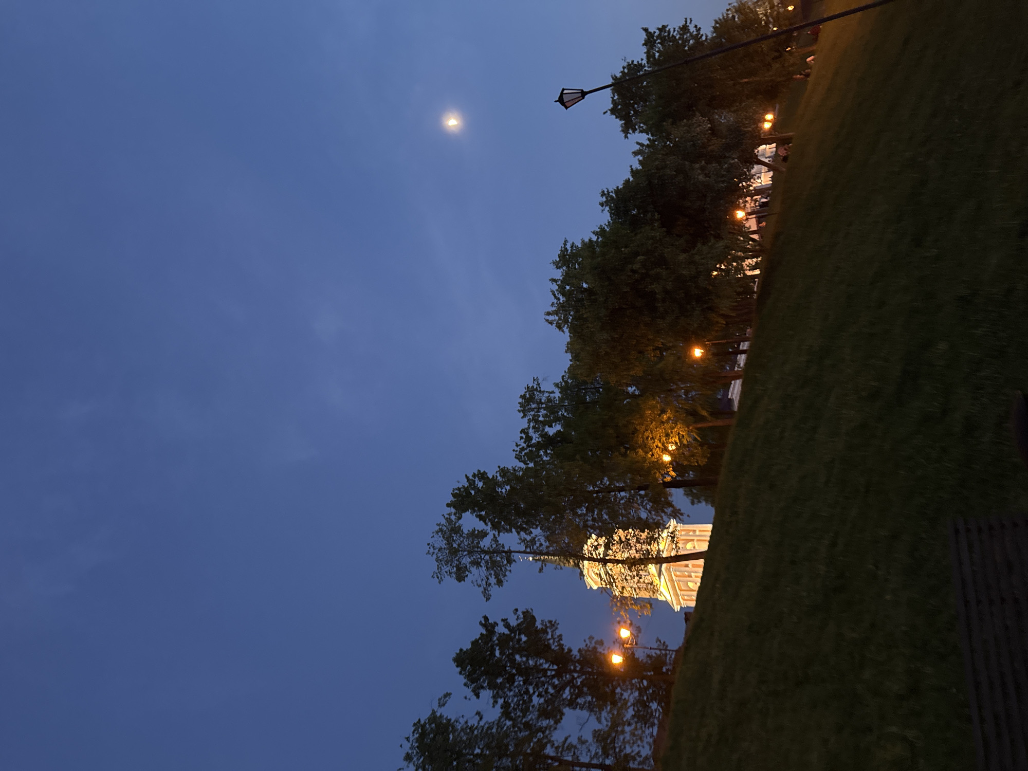 Captured at night, a low-angle shot reveals the skyline of Perm, Russia. The sky is a deep blue, with a crescent moon visible in the upper left. Trees and streetlights line the horizon, with the lights casting a warm glow. A brightly lit building with classical architecture, likely a historical landmark, stands out amidst the darkness. The foreground features a grassy slope.