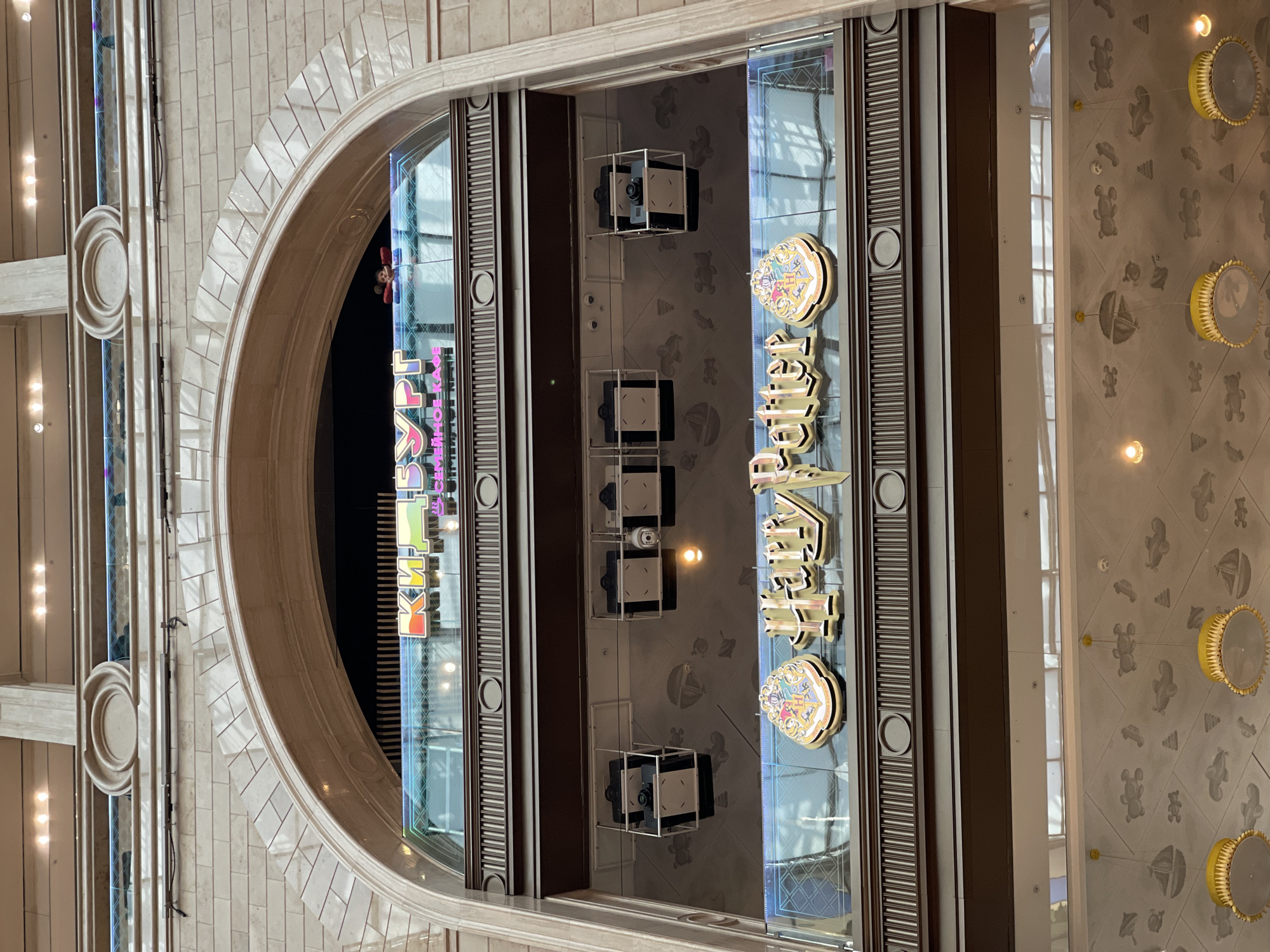 Captured from an elevated vantage point, the image presents an architectural overview of what appears to be the interior of a commercial establishment, conceivably situated in an urban environment. The vantage point allows a comprehensive view of the interior layout, encompassing the interior design elements and structural components.
The scene is dominated by the presence of signage, visible through glass panels, which suggests the presence of storefronts or establishments within the building. Architectural details, such as archways and ornate trim, are prominent features of the scene, contributing to a sense of elegance and sophistication. The overall ambiance of the space is one of commercial activity, with a focus on visual aesthetics and architectural design.
The image is a single static shot and does not show any people. The absence of temporal indicators means the exact date and time cannot be inferred from the image alone. The overall setting appears to be indoor commercial.