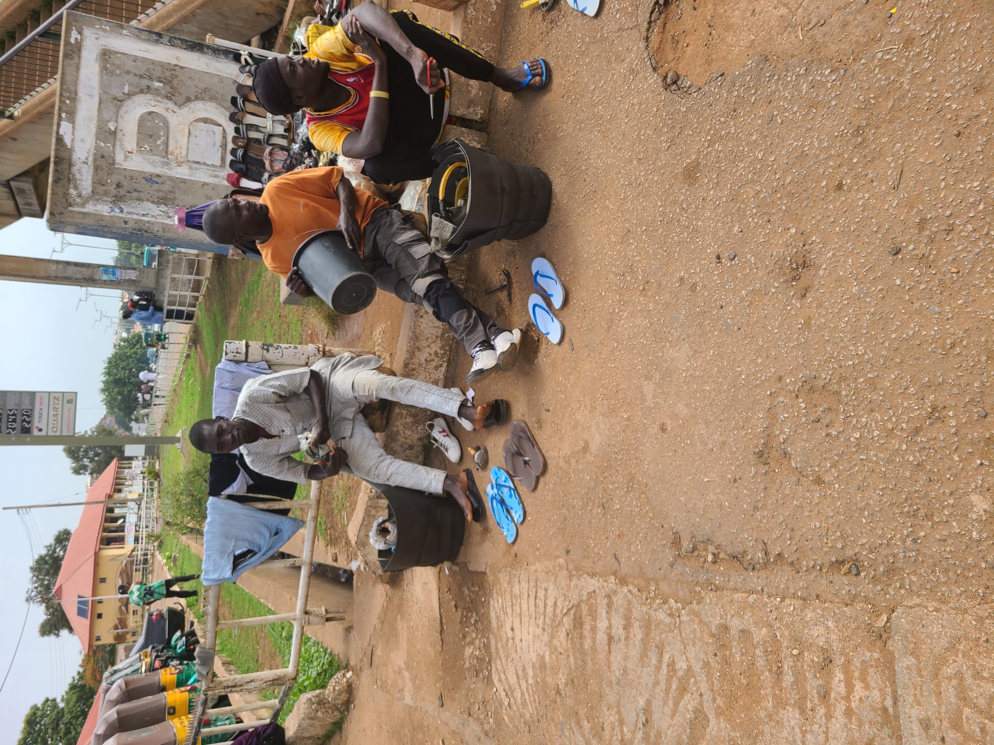 Three men are resting on a concrete ledge in an urban environment in the city of Unknown, Unknown. One man is seated with a black bucket in his lap, wearing an orange shirt and gray pants. Another man sits next to him, clad in a yellow and red basketball jersey and black pants, holding a pair of scissors. A third man sits on a ledge in the distance dressed in white; he is in front of clothes hanging on a railing. Footwear including sandals and shoes are scattered on the ground. A black bag lies on the ground. A sign in the background lists gas prices.
