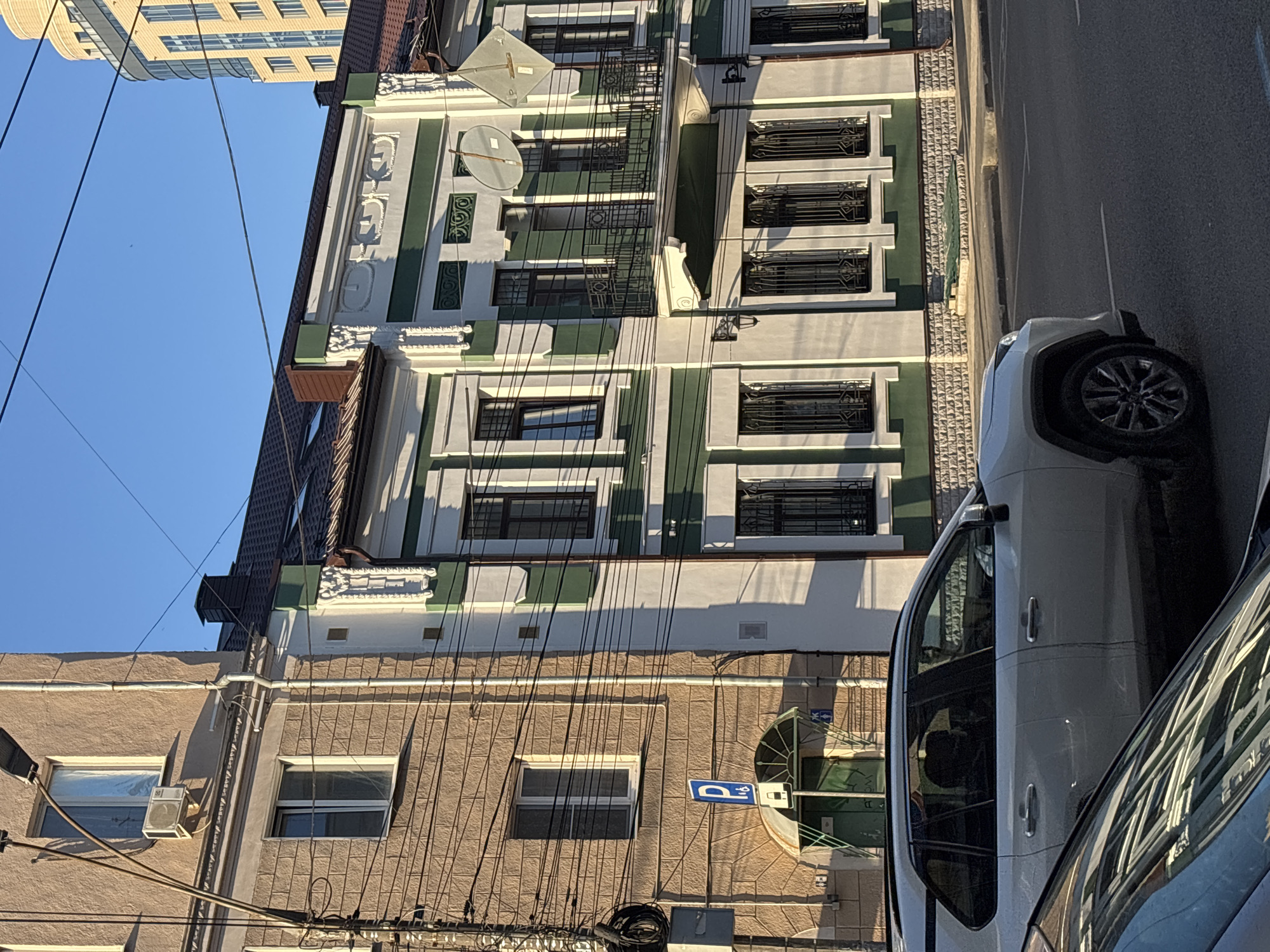 The image shows a street scene in Novaya Usman', Russia, featuring two buildings and a white SUV. The building on the left is a light-brown, multi-story structure with several windows and an air conditioning unit visible. A tall lamp post with numerous wires is attached to its facade. Adjacent to it, the building on the right is painted white with green accents, featuring decorative architectural elements, barred windows, a balcony with wrought-iron railings, and satellite dishes. A modern, light-yellow, high-rise building is visible in the background. A white SUV is parked on the street in the foreground, with another vehicle partially visible in the lower left corner of the frame, its reflection cast on the hood of the white SUV. The sky is clear and blue, indicating daylight.