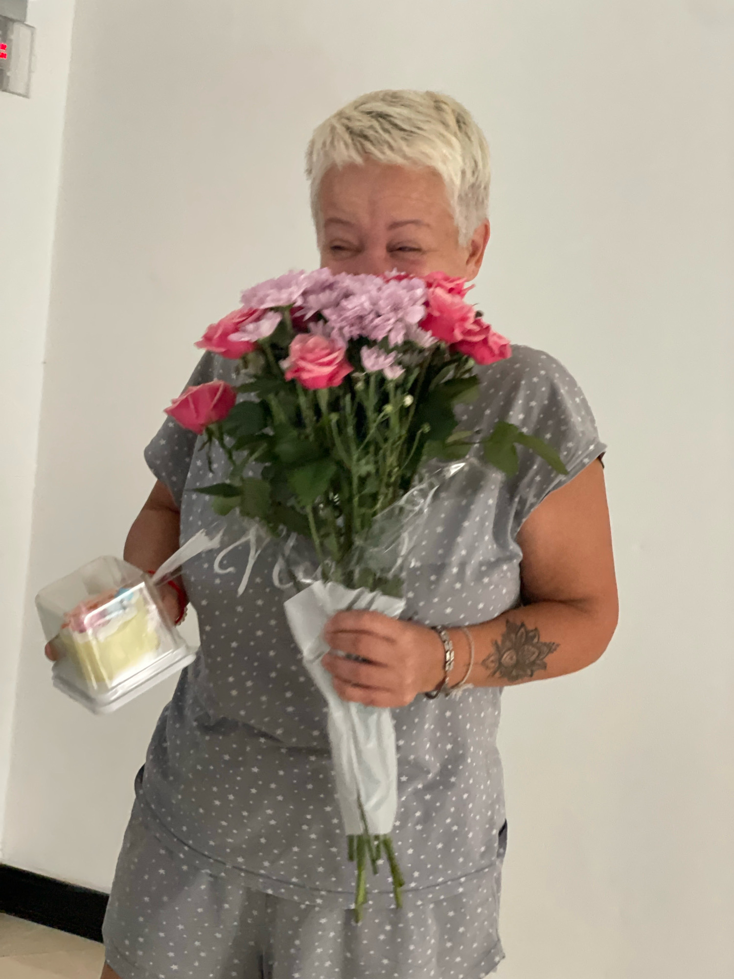 A person with short, blonde hair and fair skin is holding a bouquet of pink and purple flowers and a small slice of cake in a clear plastic container. The person is wearing a grey t-shirt with white stars and grey shorts with white stars. The person's right arm has a mandala tattoo. The background is a plain white wall. The scene is likely indoors in a residential setting, potentially in Nong Prue, Thailand.