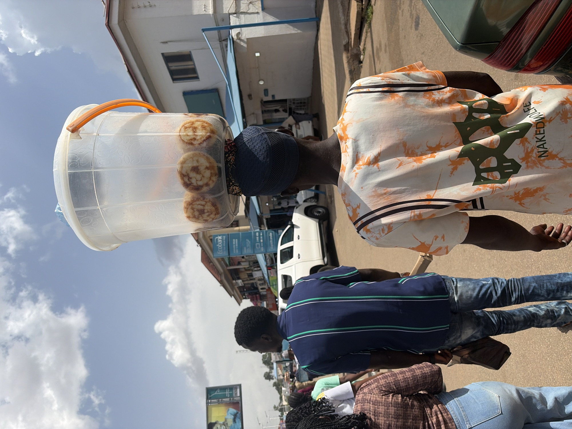 A person walks away from the camera on a paved road in Jos, Nigeria, balancing a clear plastic bucket filled with round, baked goods on their head. The bucket has an orange handle and a white lid. The person wears a blue patterned skullcap and a t-shirt with an orange, white, and black tie-dye pattern and green graphic elements on the back. Other people are visible in the background, as well as a white pickup truck, a white building with blue accents, and a billboard. The sky is cloudy.