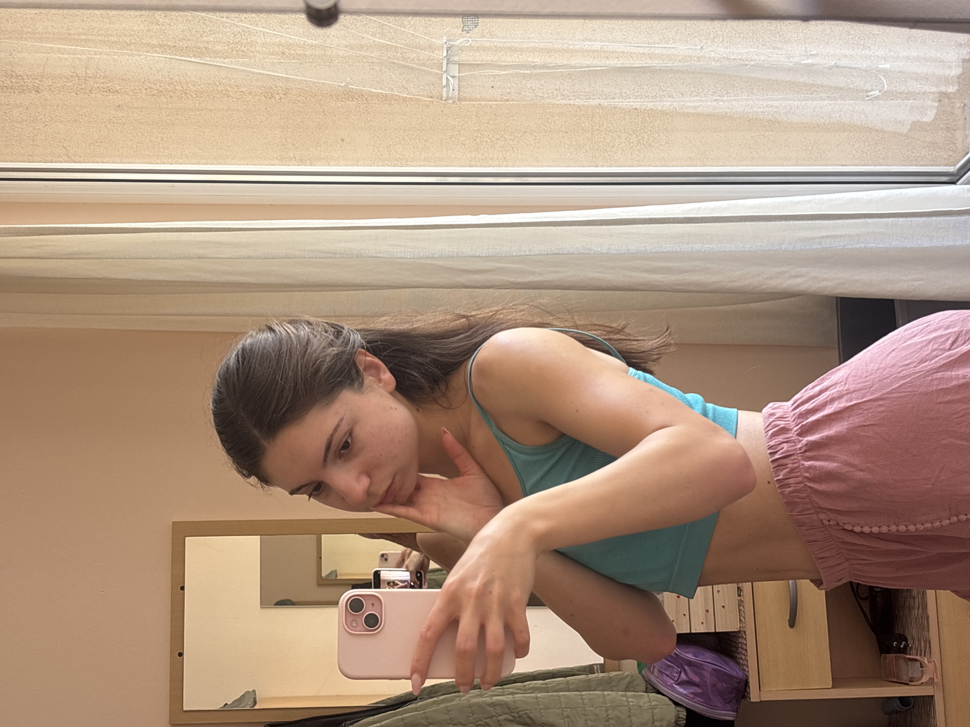 In Varna, Bulgaria, a young woman with brown hair tied back in a ponytail is taking a mirror selfie in her bedroom. She is wearing a teal tank top and pink, loose-fitting shorts. Her left hand holds a pink smartphone, and her right hand is touching her chin. The background shows a mirror reflecting the woman and her phone, a closet with a green jacket, a dresser with a purple bag, and a window with sheer curtains. The lighting suggests it is daytime.