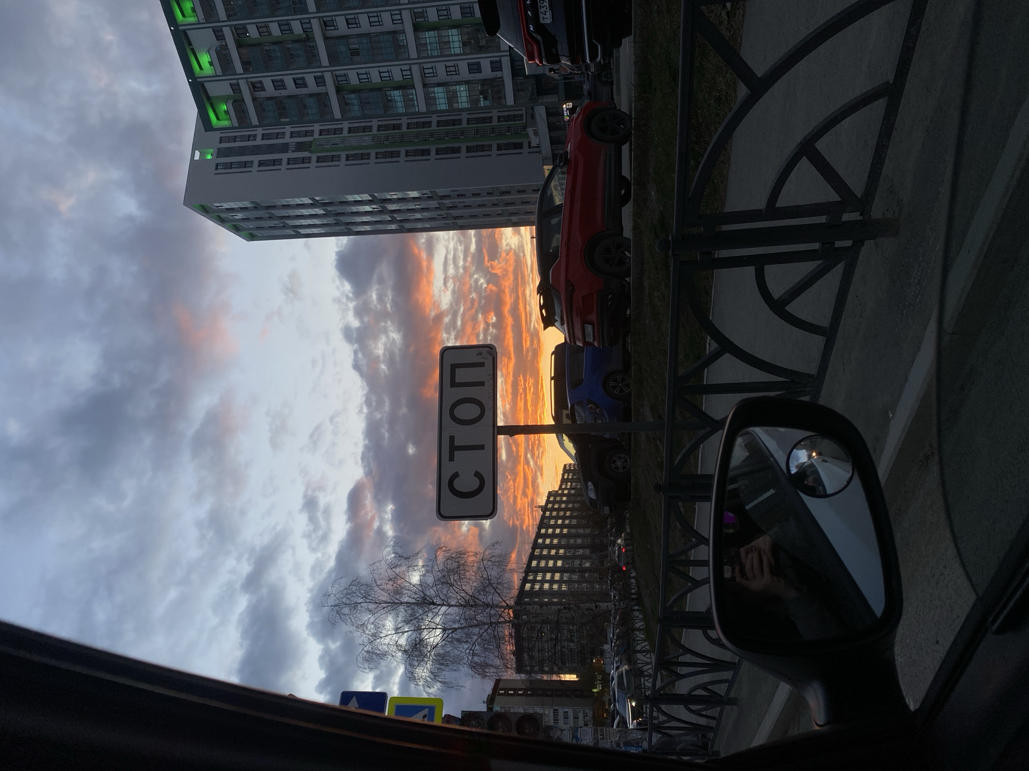 The image depicts a view from inside a car, facing outward toward a street in Yekaterinburg, Russia, at dusk or dawn. A vertical, rectangular stop sign with Cyrillic lettering is prominently visible on the left side of the frame. The sky shows a dramatic display of clouds with orange and pink hues. Two rows of buildings line the street, including a taller, modern residential building with green architectural lighting. Cars are parked along the curb. A metal fence runs along the edge of the sidewalk, adjacent to the street. The car's side mirror is visible in the foreground. A person is visible in the reflection of the side mirror, holding a phone.