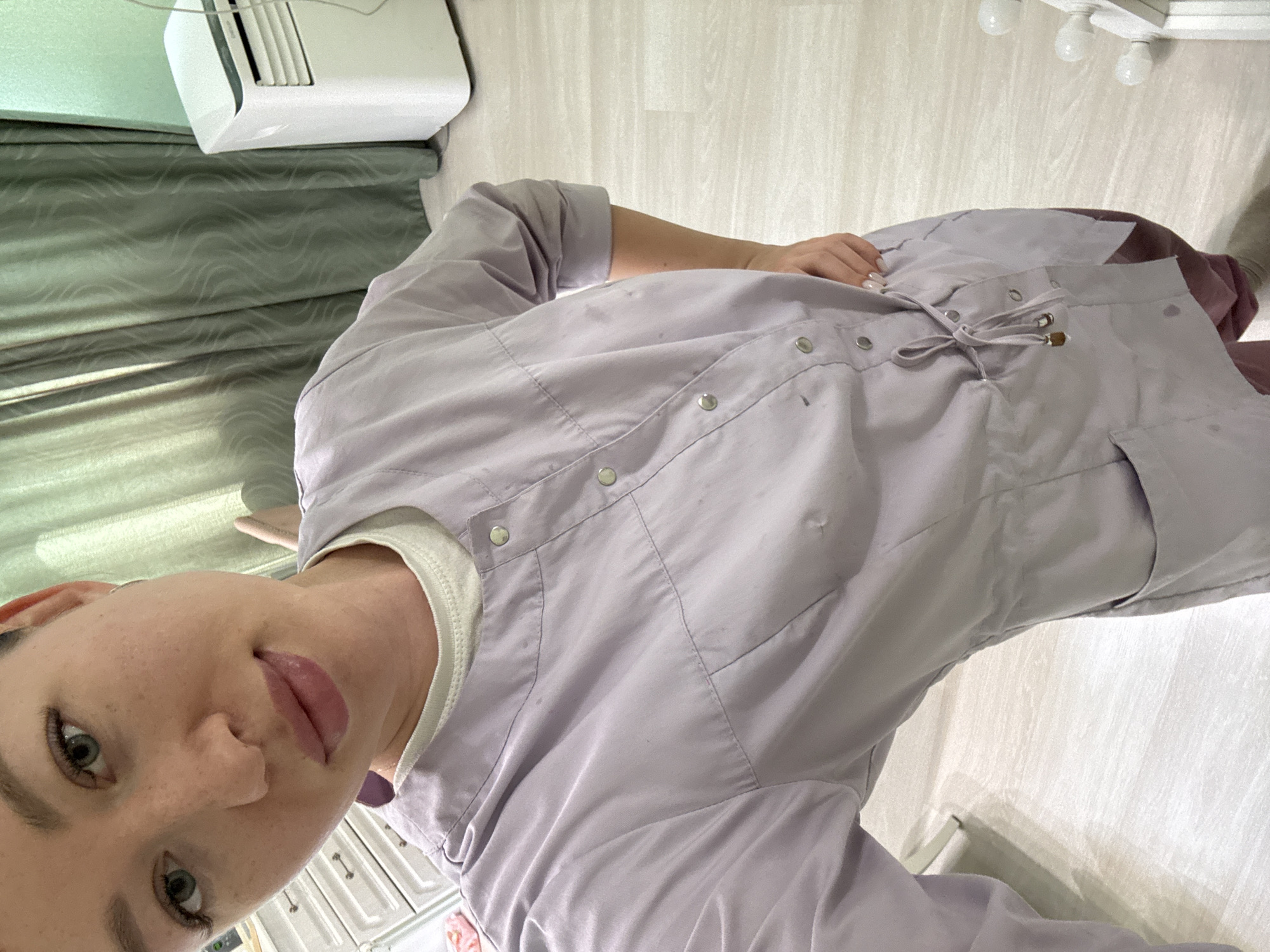 A selfie photograph taken indoors shows a young woman with fair skin, green eyes, and pink lips. She is wearing a light purple medical jacket with silver snap buttons, a white undershirt, and is holding her hand on her hip. The background includes a white mini-split air conditioner, green drapes, white cabinets, and a set of white ceiling lights. The location appears to be a medical or clinic office setting in Ферма, Russia.