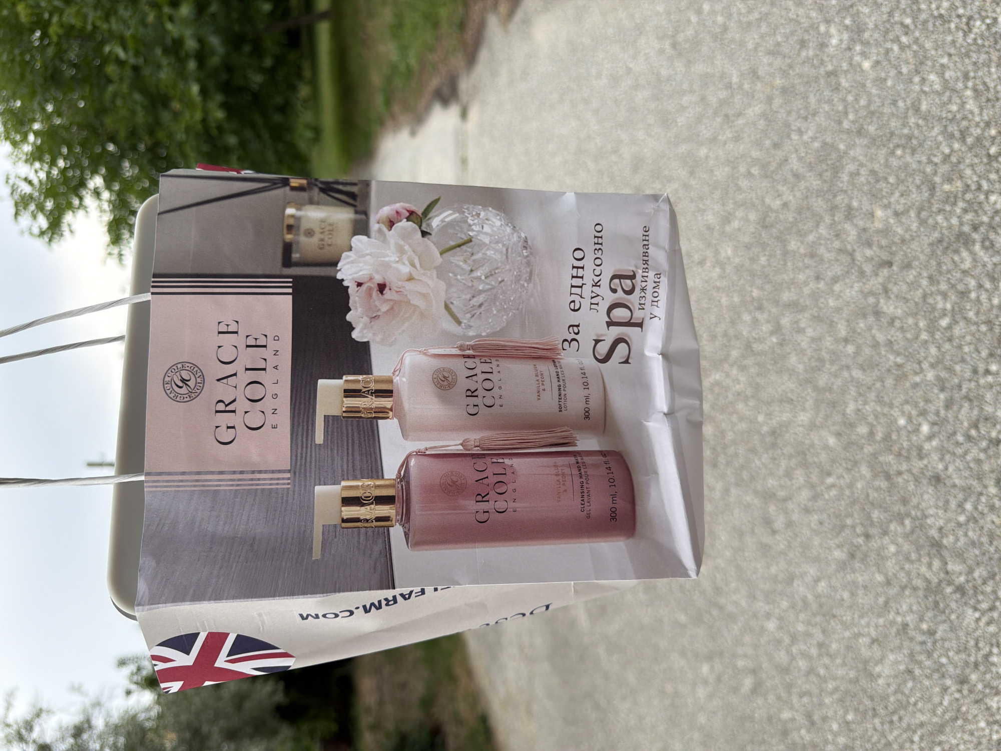 The image shows a shopping bag with a Grace Cole England advertisement on it, held in front of an outdoor background. The advertisement features two pink pump bottles of hand wash and lotion, along with a small vase of pink peonies and a candle. Text on the bag includes "GRACE COLE ENGLAND," product names, and Bulgarian text which translates to "For an exquisite Spa experience at home." The background appears to be a paved walkway in a park-like setting, with green trees and foliage visible. There is no visible indication of the specific location within Pazardzhik, Bulgaria. The image is taken from a slightly low angle, focusing on the shopping bag which is the main subject.