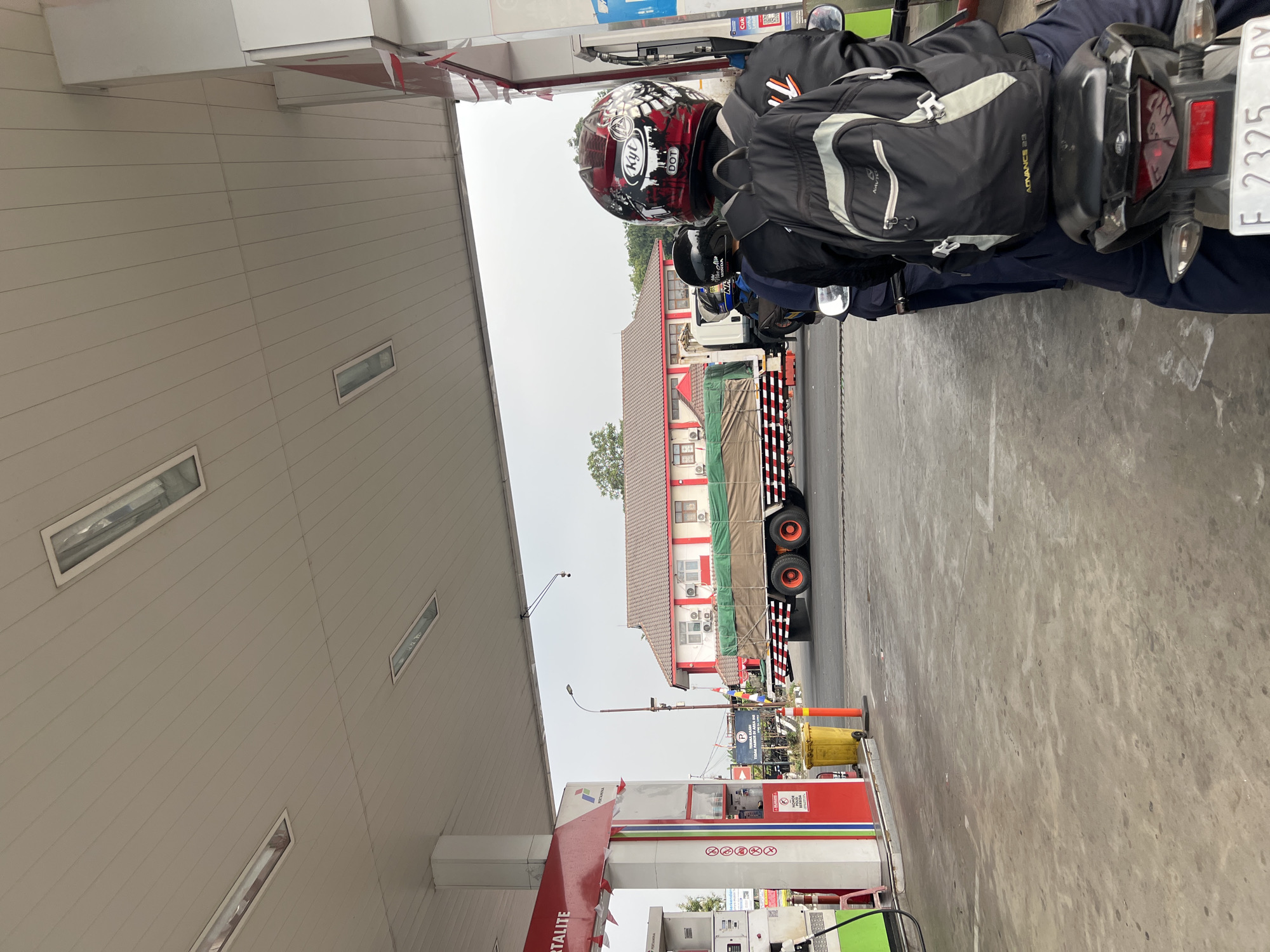 The image depicts a gasoline station in Gunung Putri, Indonesia, as evidenced by the "PERTAMINA" branding on the fuel pumps and the visible license plate "F 2325 RY," which is consistent with Indonesian vehicle registration. The scene is viewed from the perspective of someone on a motorcycle, waiting in line at a gas pump. In the foreground, several individuals on motorcycles are lined up. The motorcycle in the immediate foreground has a rider wearing a black jacket and a backpack. Behind them, other riders are visible, also wearing helmets.
The gas station canopy is visible overhead, with fluorescent lighting. To the left, a Pertamina fuel pump is prominent, with red, white, and green accents. A yellow trash bin is situated near the pump. In the mid-ground, a large truck with a green tarp-covered cargo is stopped on the road, partially obscured by the canopy. The truck has a distinctive red and white striped bumper. Behind the truck, a building with a red-tiled roof, white walls, and red trim is visible. The sky is overcast. The ground is paved with concrete and shows some stains and markings. The overall impression is of a typical daytime scene at a busy roadside gasoline station.