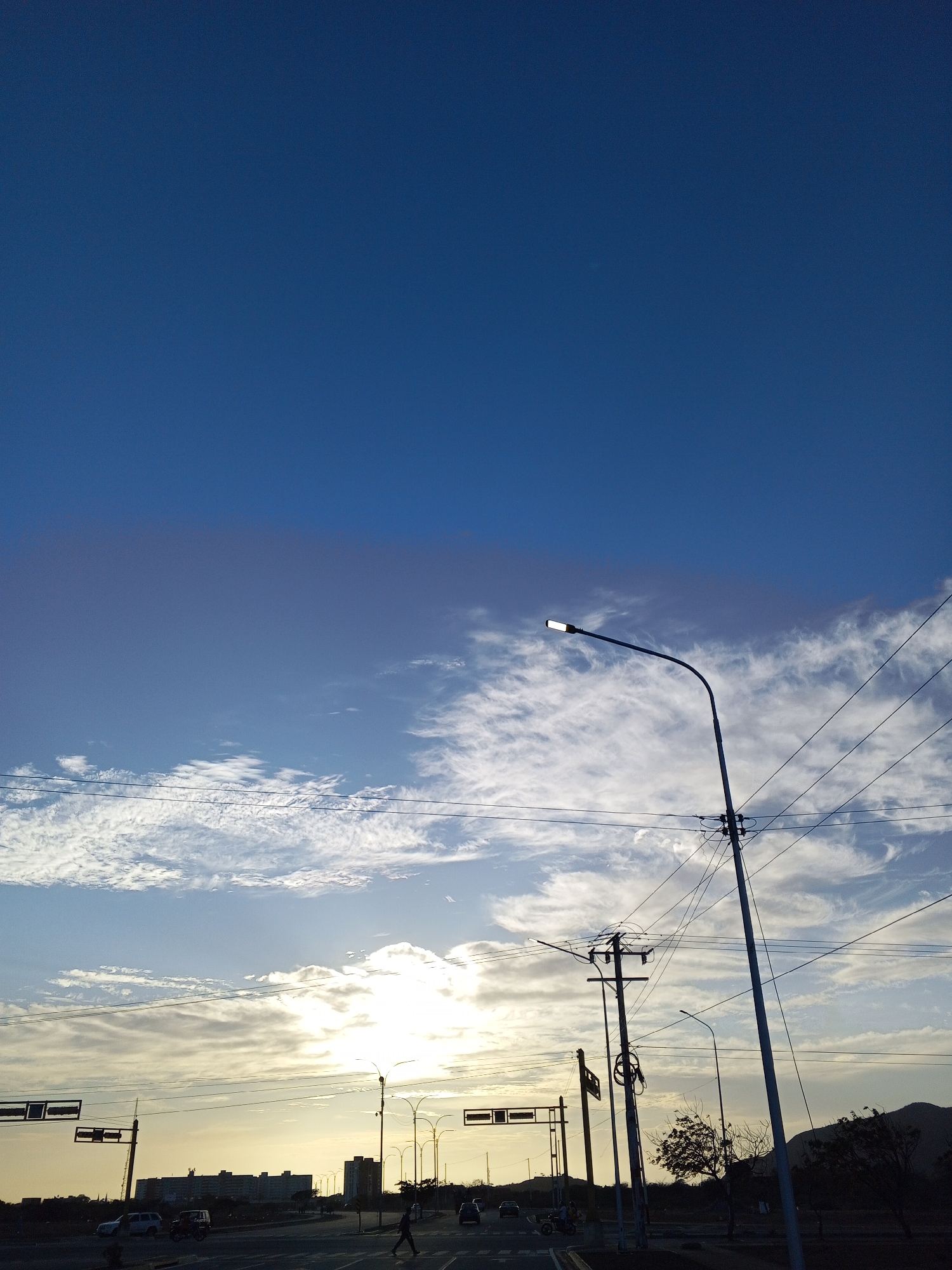 The image captures a streetscape in Altagracia, Venezuela, at what appears to be dusk. The sky transitions from a deep blue at the top of the frame to a band of wispy clouds near the horizon, where the sun is setting. Overhead power lines crisscross the scene, connected to a series of utility poles and a modern street lamp that is illuminated.
Below the horizon line, a multi-lane road is visible, with cars and a single pedestrian crossing. Traffic signals and buildings are silhouetted against the bright light of the setting sun. In the distance, a low mountain range can be seen.