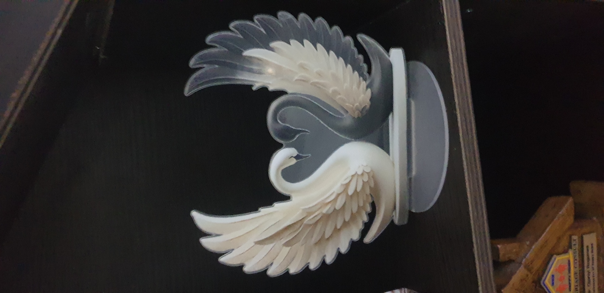 The image displays a decorative figurine of two swans forming a heart shape, positioned on a shelf. The swan on the left is white with cream-colored wings, while the swan on the right is dark grey with translucent greyish wings. Both swans have their necks curved inward to create the upper outline of a heart. They stand on a light grey, multi-layered base that exhibits a slight translucency at its lower edge, suggesting a reflective surface underneath. The background behind the figurine is uniformly dark, indicating a dark wall or the interior of a display case. A wooden shelf or surface is visible beneath the figurine, running across the bottom portion of the frame, showing faint horizontal lines consistent with wood grain. The lighting appears even, highlighting the details of the swans and their base. The specific location of Barden Yana, Nigeria, is not discernible from the visual information provided within the image.
