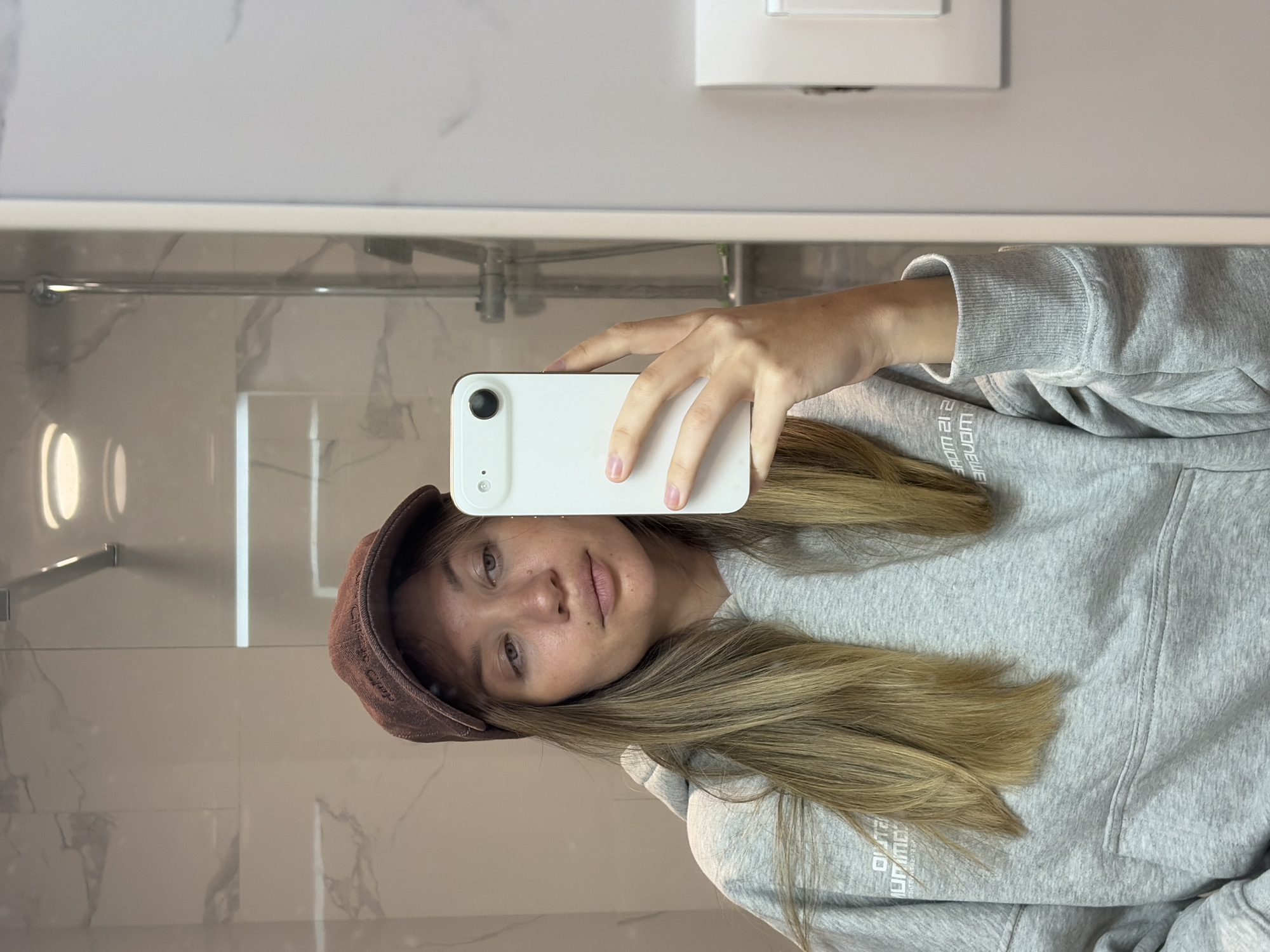A young woman is taking a selfie in what appears to be a bathroom. She is wearing a brown cap and a gray hoodie with white lettering. She is holding a white smartphone up to her face. The background features a marble wall, a shower fixture, and a light switch. The location is Unknown, Unknown.