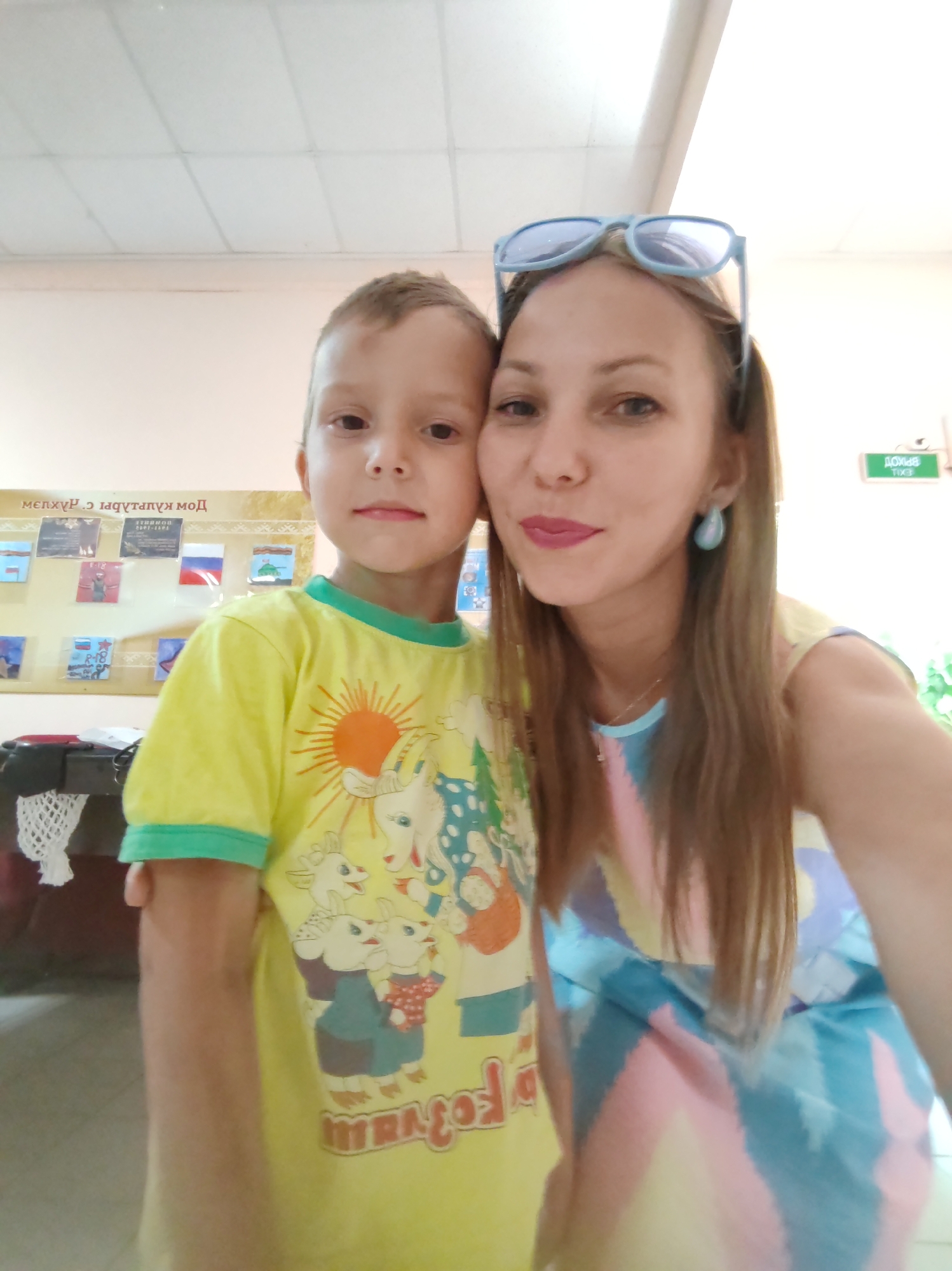 A young boy and a woman are posing for a selfie in an indoor environment that appears to be a classroom or community center. The boy, on the left, is wearing a bright yellow t-shirt with a colorful cartoon print featuring animals and a sun. The woman, on the right, is wearing a dress with a pastel, abstract striped pattern and light blue sunglasses on top of her head. She has light-colored earrings. The background shows a bulletin board with various small pictures and text, and a green exit sign with Cyrillic lettering reading "ВЫХОД" (Exit). The overall lighting suggests an interior setting with artificial light.
