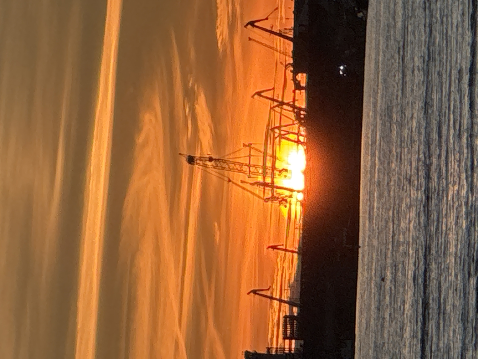This is a horizontal, medium shot of a harbor scene at sunset in Tallinn, Estonia. The sky is a vibrant orange and yellow, with wispy clouds stretching across the frame. The sun is low on the horizon, casting a warm glow over the scene and is partially obscured by several cranes. These cranes are silhouetted against the bright sky and appear to be part of a port or industrial area. In the foreground, there is calm water with gentle ripples. The overall impression is one of industrial activity set against a beautiful natural backdrop.