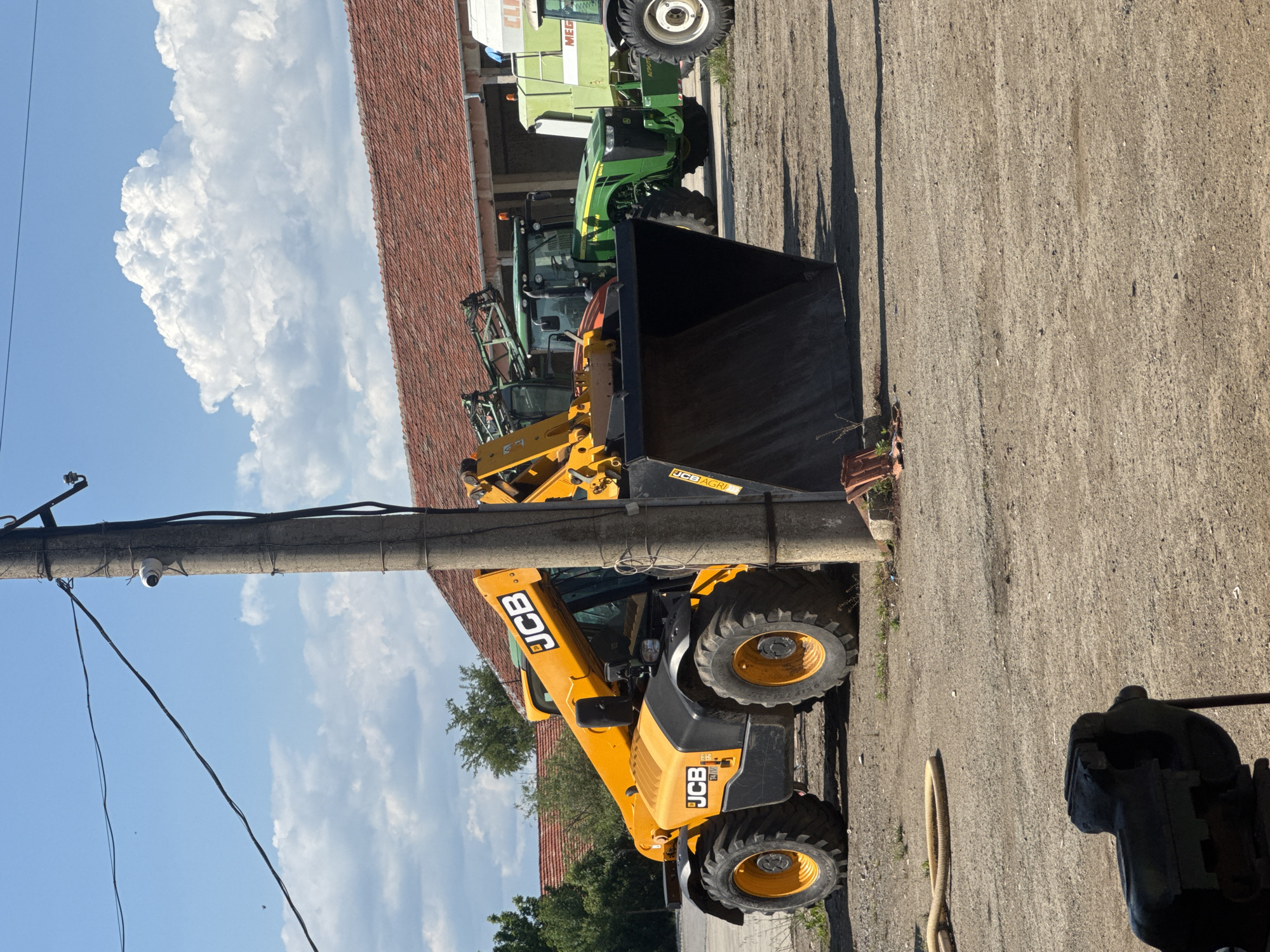 The image depicts a bright yellow JCB telescopic handler parked on a paved surface alongside a row of other agricultural vehicles, including a green John Deere tractor and a CLAAS combine harvester. The handler is positioned close to a concrete utility pole with attached electrical wires and a security camera. The background shows a building with a red-tiled roof and a partially visible treeline. The sky is blue with puffy white clouds. A dark metal object of indiscernible form is present in the bottom right corner. Based on the image context, it is plausible that this scene is located in an agricultural area, potentially in Sarnevo, Bulgaria, or in a similar setting where agricultural machinery is commonly used and stored. There are no people visible in the frame.