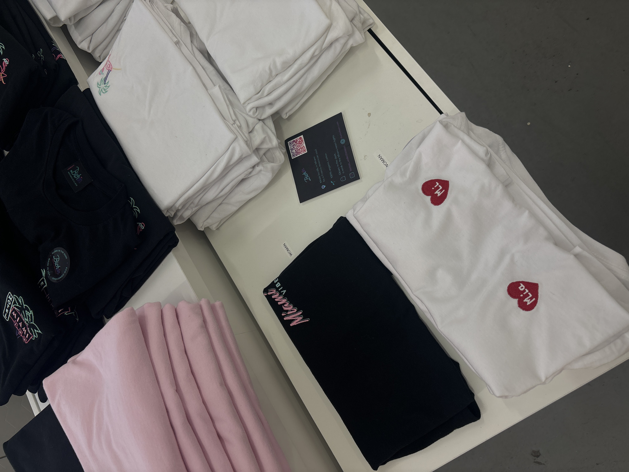 The image presents a selection of folded clothing items, presumably shirts, neatly arranged on a white shelving unit. The clothing displays various designs and colors, including black shirts with "Miami" and neon-themed graphics, white shirts with a palm tree design and heart-shaped logos with the name "Mia" embroidered, and a stack of pink shirts.
A black card with a QR code and the brand name "Bichon" is placed among the shirts, featuring marketing details about the locally made, organic cotton clothing brand from Miami. The image suggests these clothing items might be part of a retail display showcasing Miami-themed apparel.