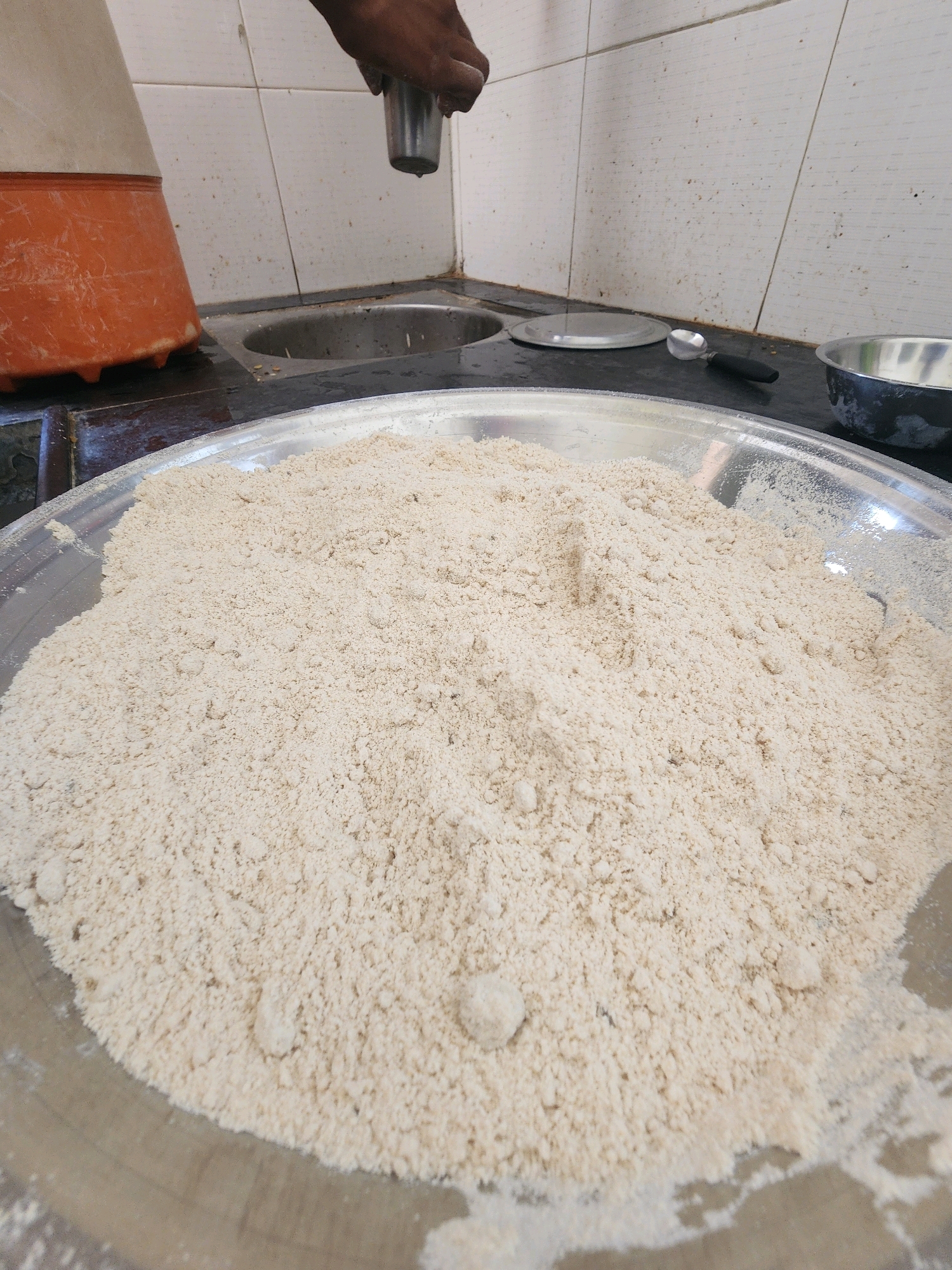 A medium shot shows a large metal basin filled with a mound of light-brown flour. In the background, a person's hand is visible holding a small stainless steel cup, from which a single drop of liquid is falling. To the left, an orange plastic container with a white lid is partially visible. The countertop is black, with a stainless steel sink to the right of the orange container. Behind the sink, white tiled walls are present. A small serving spoon with a black handle rests on the countertop near a stainless steel bowl on the right. The scene appears to be a kitchen or food preparation area, likely in Mahesana, India, given the context.