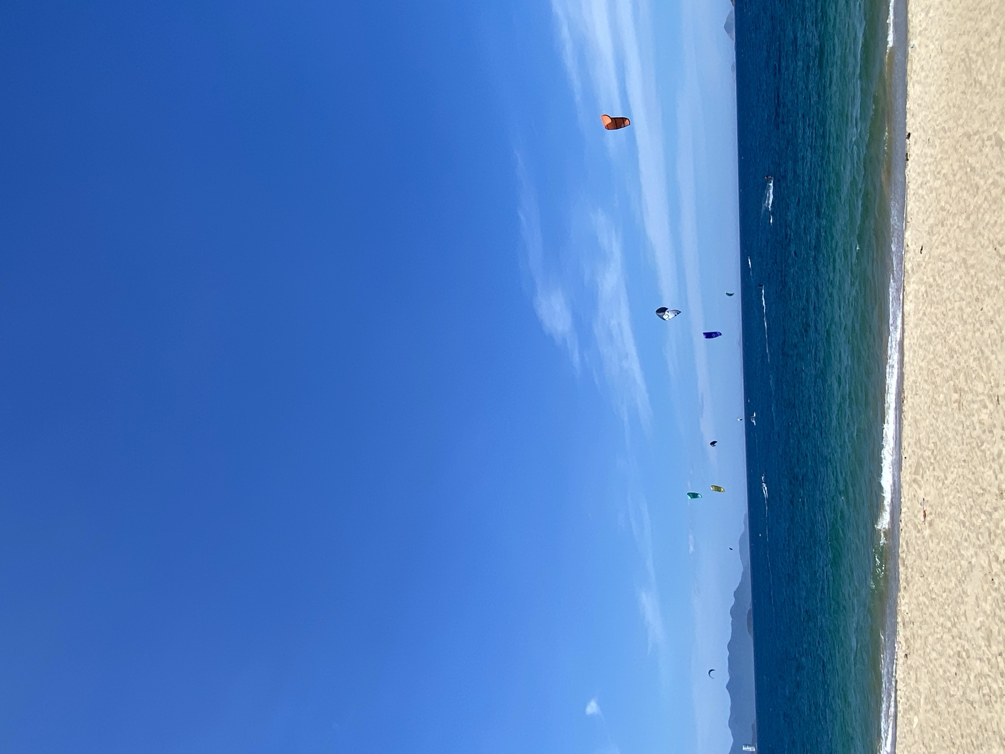 The image depicts a bright, sunny day at a beach in Nha Trang, Vietnam. The foreground is a wide expanse of pale sand with gentle waves lapping at the shore. The ocean is a vibrant blue, transitioning to turquoise near the shore, with a few whitecaps indicating a gentle breeze. Several kitesurfers are visible in the water, their colorful kites dotting the horizon against the clear blue sky. In the distance, a hazy mountain range forms the backdrop, with some buildings visible on the left side of the image. The sky is a deep, cloudless blue, with a few wispy white clouds scattered across it. The scene conveys a sense of activity and recreation in a coastal environment.