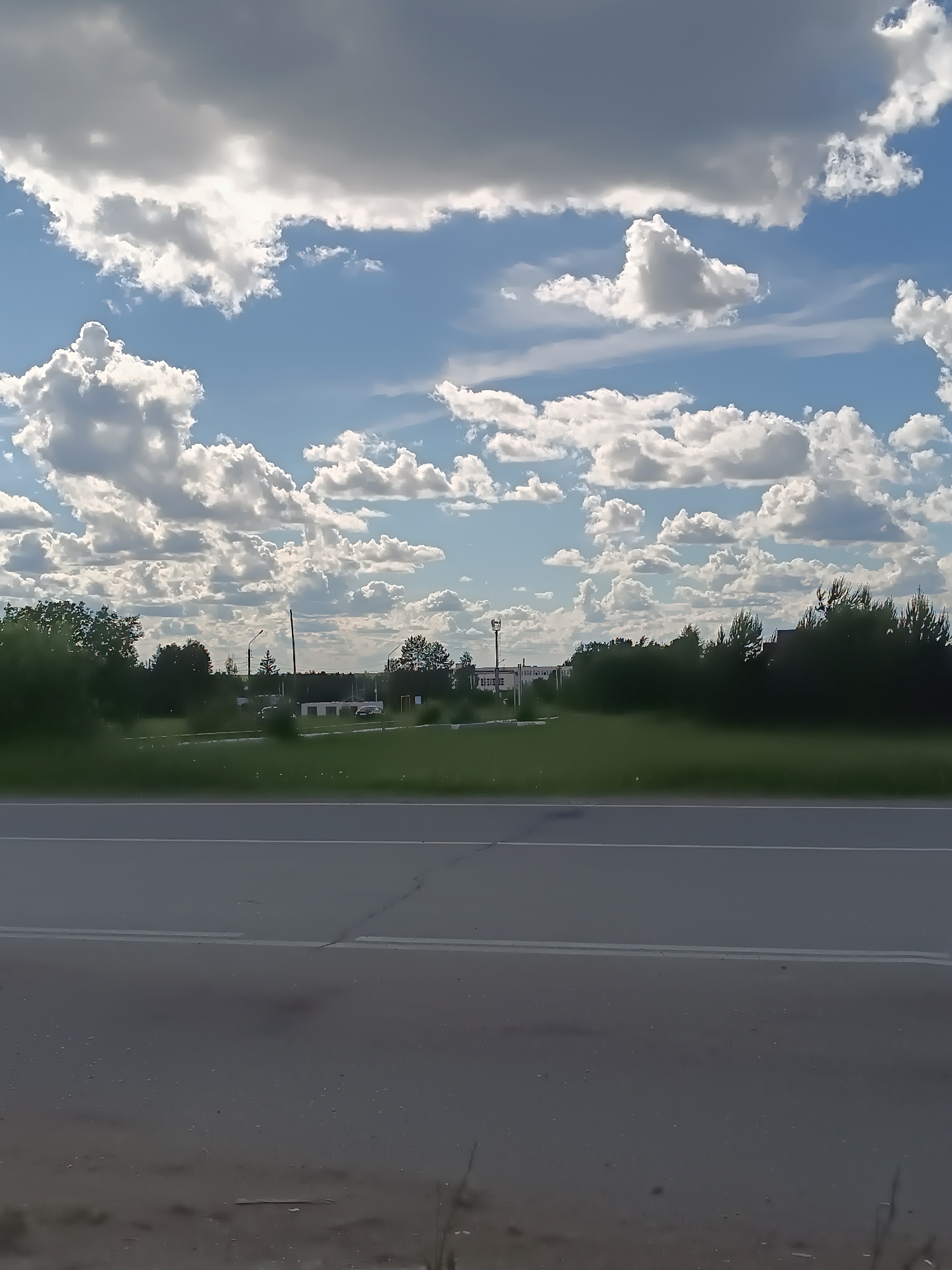 The image shows an asphalt road with two white lane markings. Beyond the road, there is a green grassy area with scattered trees and shrubs. In the background, there are buildings and structures. The sky is blue with numerous white, fluffy clouds. The location appears to be a rural or suburban area, possibly on the outskirts of a city.