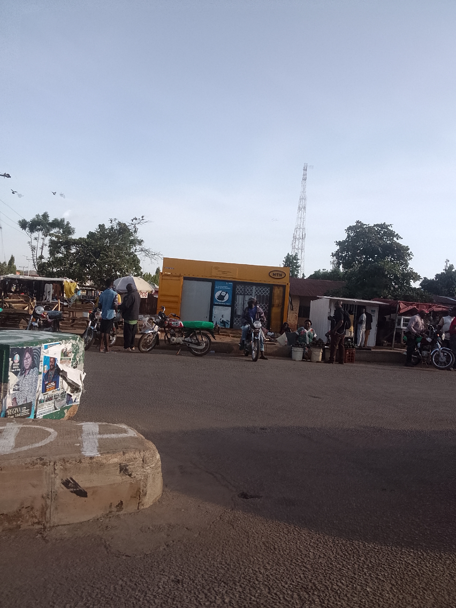 The image depicts a street scene in Bukuru, Nigeria. The photograph is taken at ground level. In the foreground, a concrete barrier is positioned on the left with posters affixed to it. The street is paved.
In the middle ground, several people are seen standing and sitting along the side of the road. Motorcycles are parked nearby. A yellow building with "MTN" signage is visible, suggesting it could be a telecommunications service provider outlet. There are also stalls and smaller structures that appear to be informal businesses or shops.
In the background, a tall telecommunications tower rises into the sky. Trees dot the landscape, and the sky is light blue with some clouds.