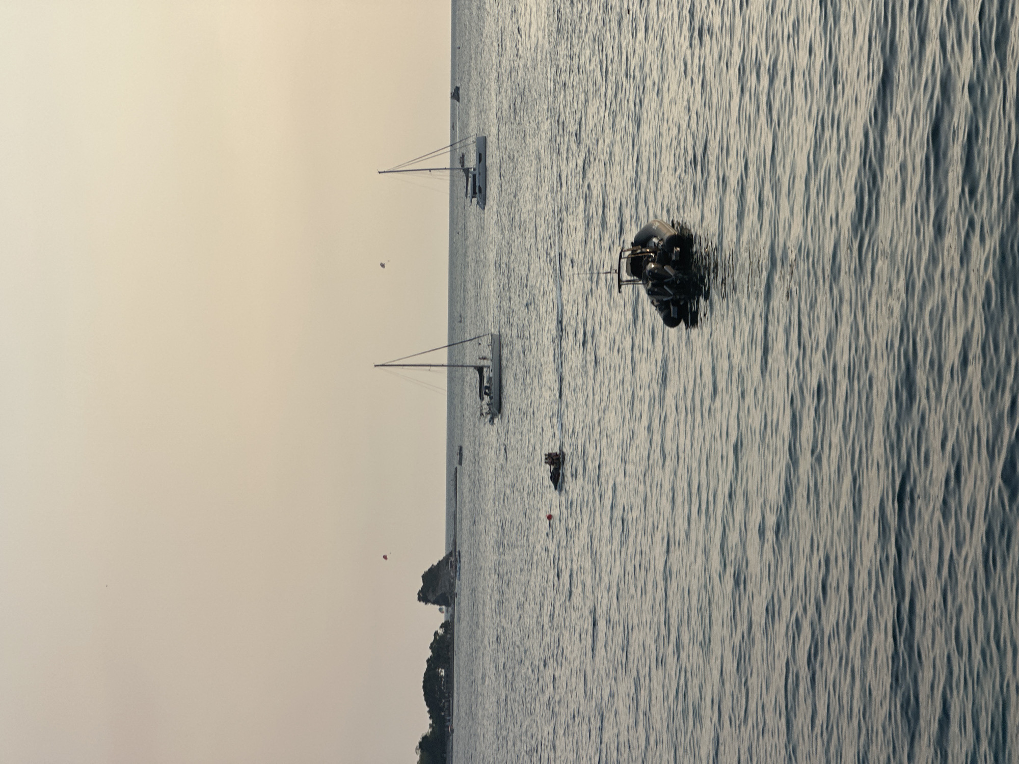 A serene, muted sunset over the Adriatic Sea near Budva, Montenegro, frames a tranquil scene with several vessels and a distant coastline. In the foreground, a dark, inflatable rigid-hulled boat with twin outboard motors is anchored, its polished surfaces reflecting the dim light. Further out, a person is riding a jet ski, leaving a small wake, with a red buoy nearby. Two sailboats with their masts reaching towards the pale sky are positioned in the mid-ground. One sailboat, a catamaran, is a bit closer and to the right, while a monohull sailboat is to its left. In the distance, a small, rocky island covered with trees, likely part of the Budva archipelago, is visible, with structures and smaller boats dotting the shoreline. The sky is a soft gradient of pale orange and cream, with a few faint specks that might be birds or distant aircraft. The sea is calm, with subtle ripples that catch the fading light.