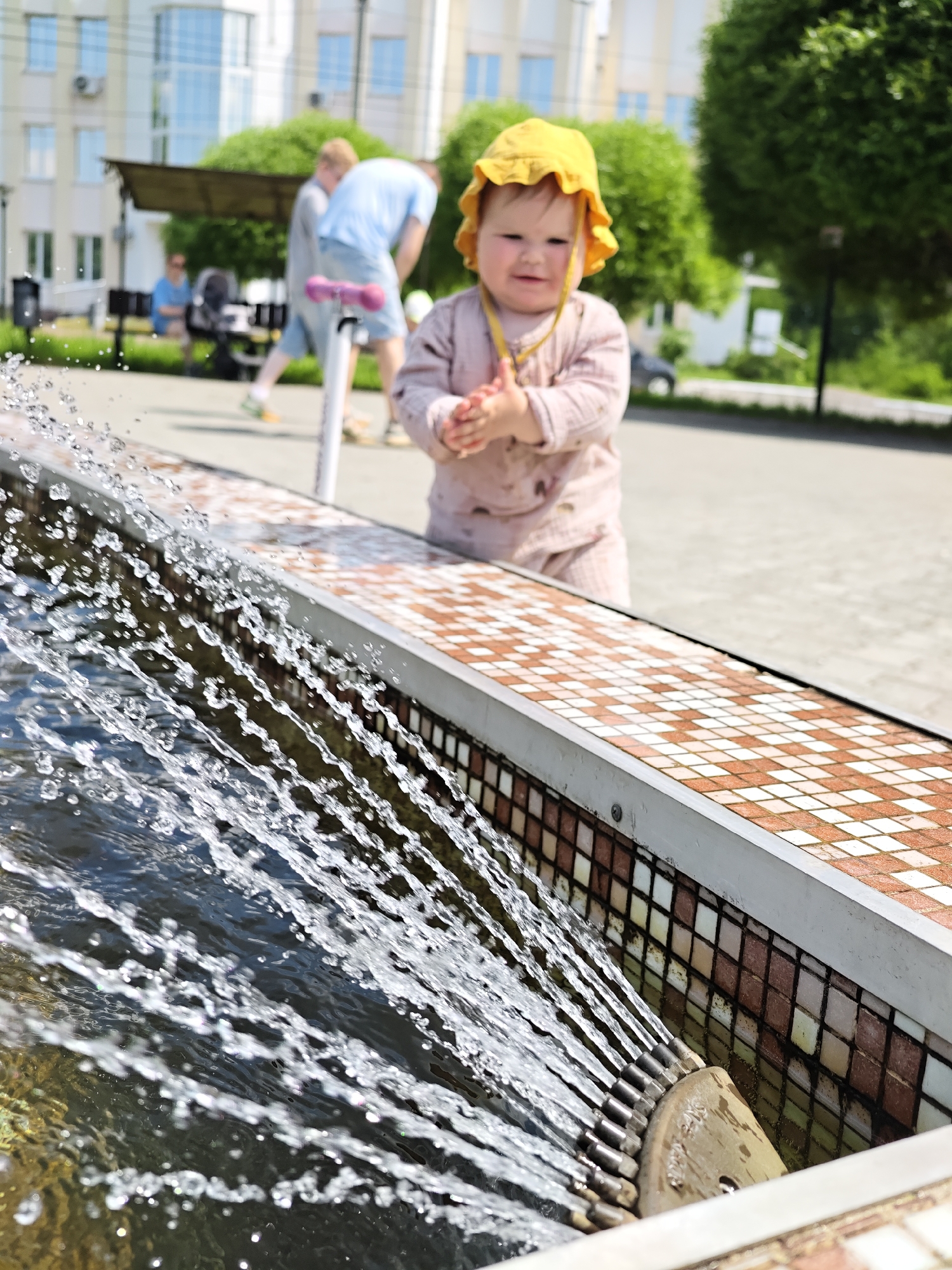 In the city of Chaikovsky, Russia, on a bright, sunny day, a young child wearing a yellow sun hat and a pink romper is standing near a fountain. The child is clapping their hands, looking towards the camera with a smile. In the background, a person in a blue shirt and shorts is bending down near a scooter, and another person is seated on a bench. The fountain is constructed with a tiled edge, featuring a pattern of small squares in shades of white and reddish-brown. Water sprays from the fountain, creating a dynamic visual element in the foreground. The environment appears to be an outdoor public space, possibly a park or a plaza, with buildings and trees visible in the distance.