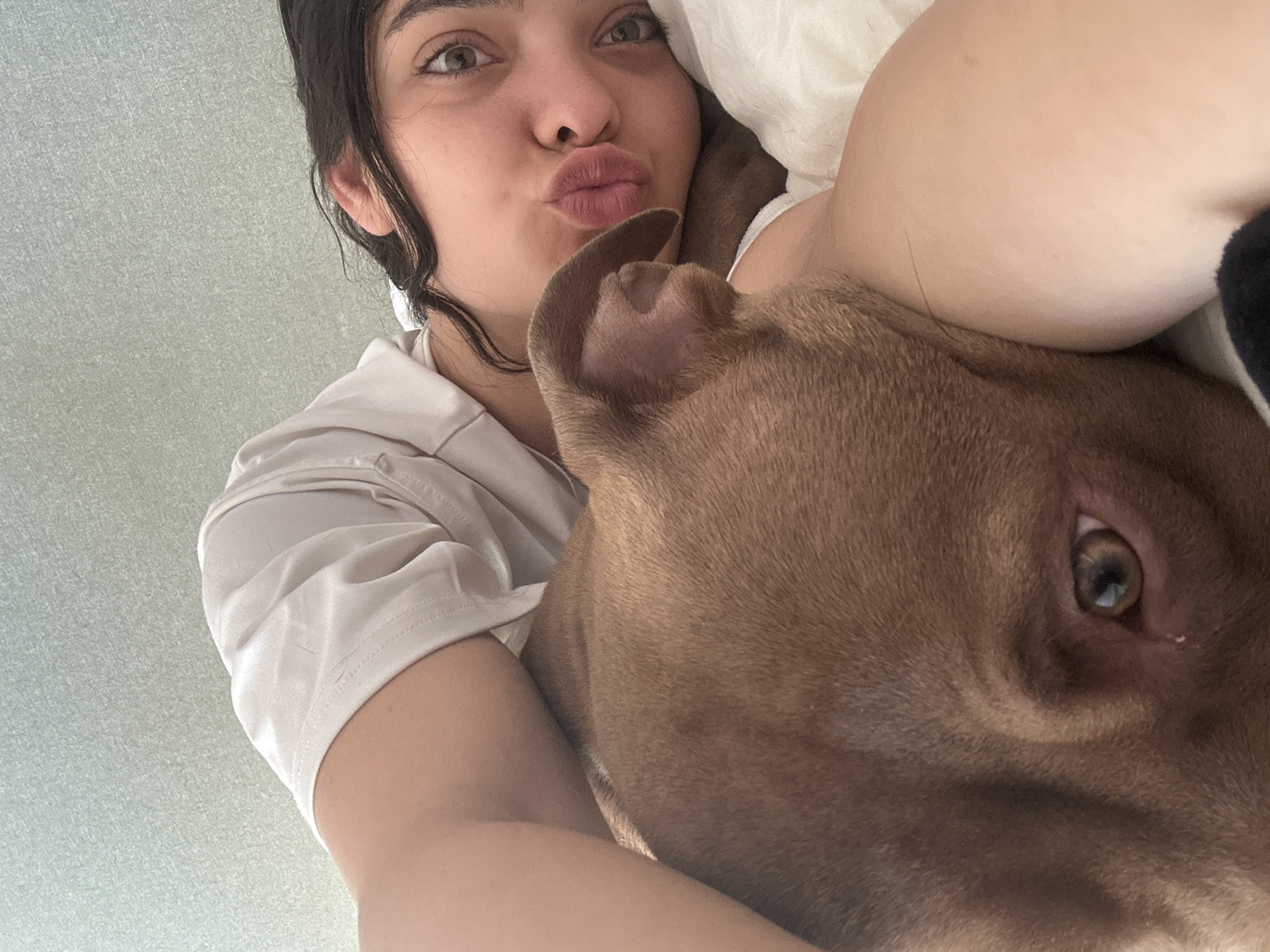 A close-up, selfie-style shot shows a young person lying in bed with their head resting on their arm. A brown dog is positioned in front of them, its head near the person's face. The person is wearing a white t-shirt and has dark hair and brown eyes. Their lips are pursed. The dog has a light brown coat and one ear is visible. They are both lying on a white bed with a light green textured wall in the background. The scene appears to be indoors, likely a bedroom. The location is the city of Malevo, Bulgaria.