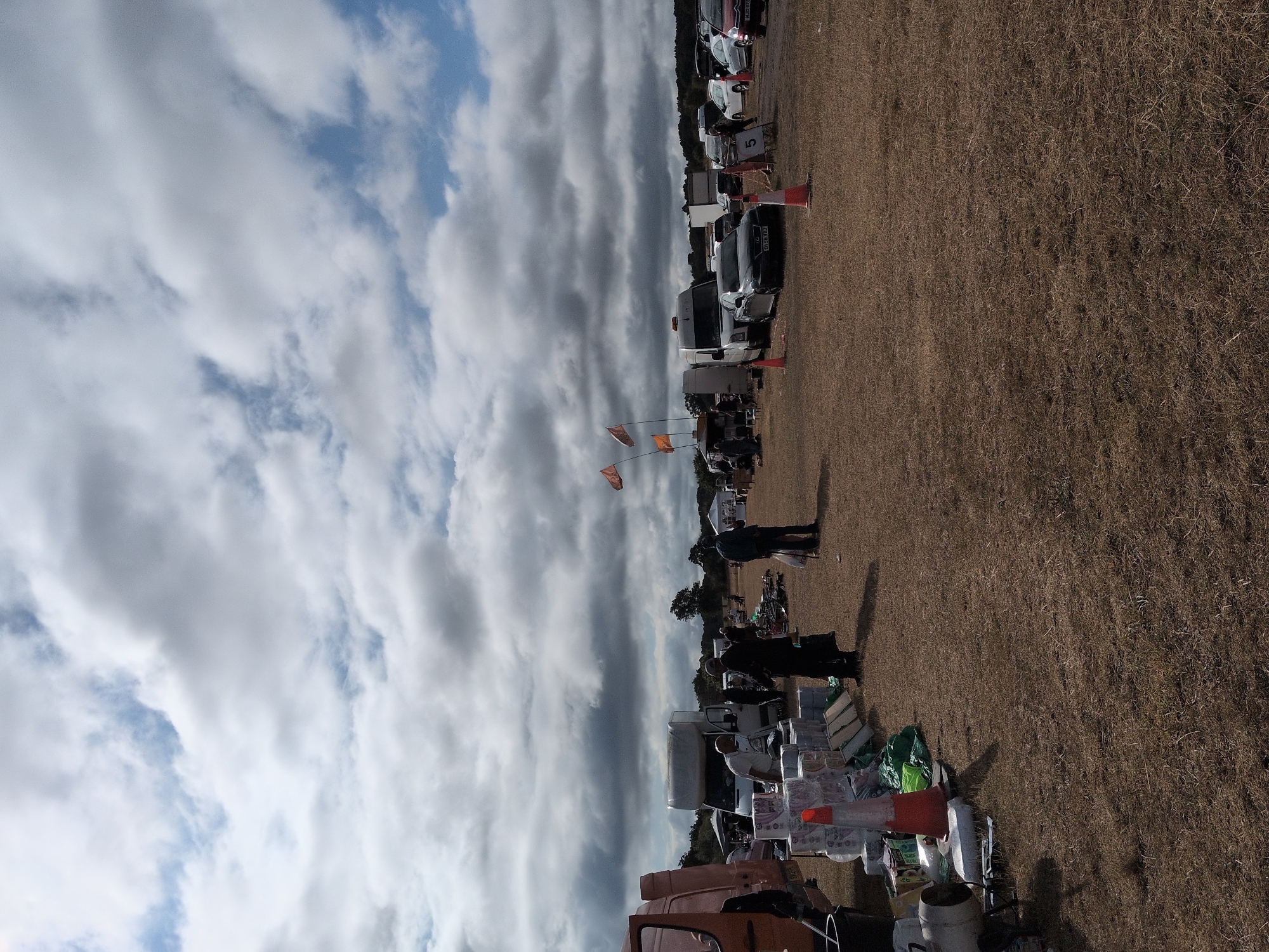 The image depicts an outdoor market or car boot sale on a dry, grassy field under a cloudy sky. In the foreground, there are several stacked items, including what appear to be rolls of toilet paper, a black bucket, and a large orange traffic cone. A person in a black jacket and trousers stands near the toilet paper stacks, facing away from the camera. To the left, part of an orange van is visible.
In the middle ground, several vehicles are parked, including white vans and cars. People are scattered throughout the scene, some standing near the vehicles and others interacting with stalls. Two orange flags with what appear to be symbols on them are visible on poles to the left of the frame. A sign with the number "5" is visible in the middle right of the image.
The environment is a flat, open field with sparse, dry grass. In the background, trees and a distant tree line are visible. The sky is heavily clouded with patches of blue sky peeking through. The overall impression is of a busy, casual outdoor event taking place during daylight hours. The location is identified as South Staffordshire, United Kingdom, suggesting this is a local community event.